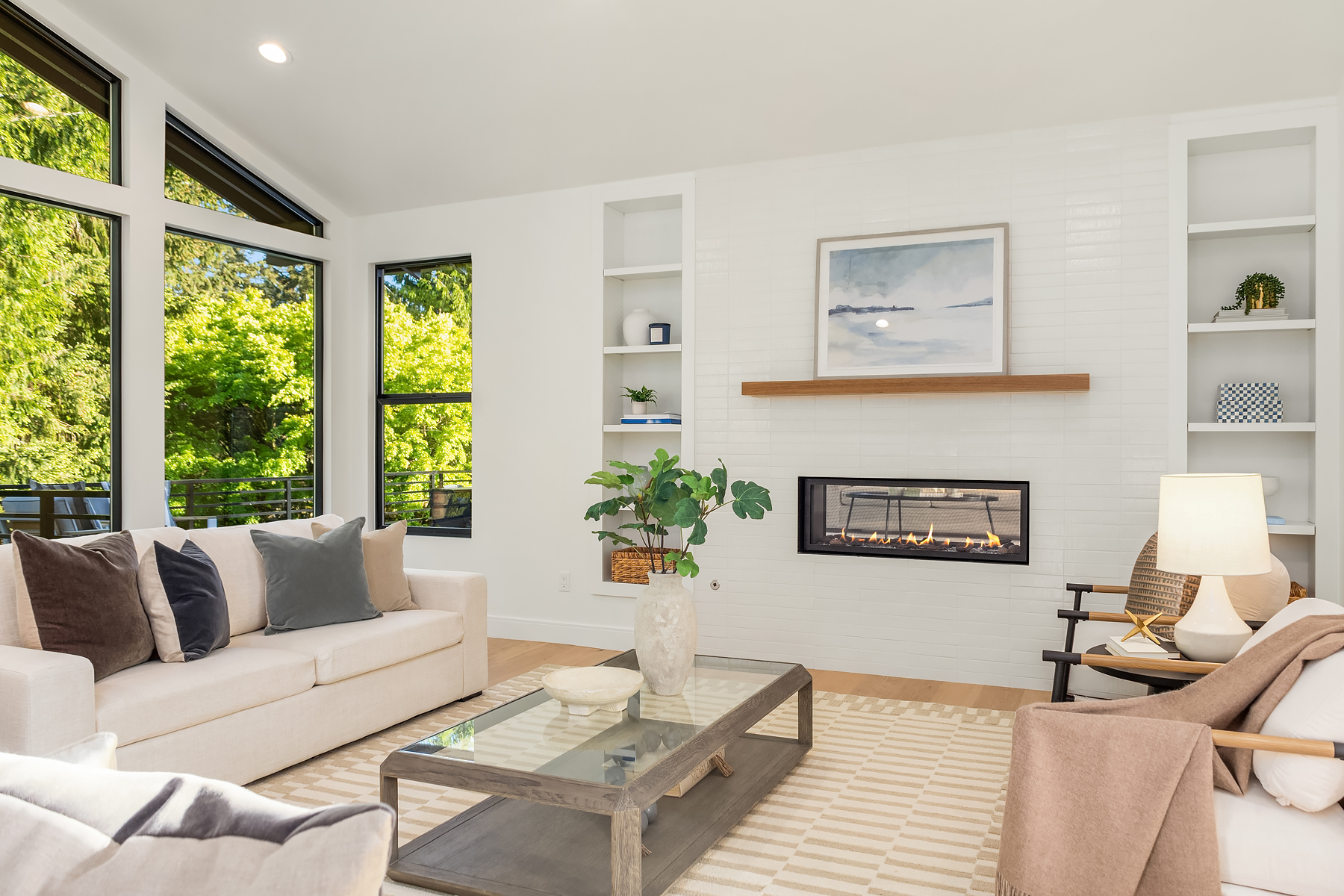 Bright, modern living room with large windows, white walls, a beige sofa, glass coffee table, built-in shelves, a fireplace, potted plant, and neutral-toned decor. Sunlight streams in from outside.