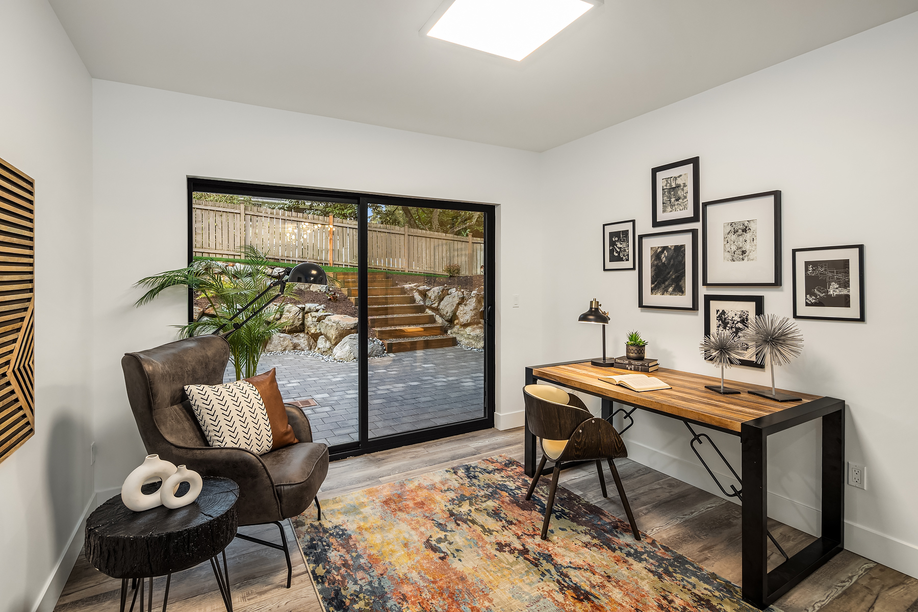 Modern home office with a large window overlooking a patio and garden, featuring a wooden desk, two chairs, a patterned rug, wall art, and plants. Natural light fills the room, creating a cozy, inviting atmosphere.