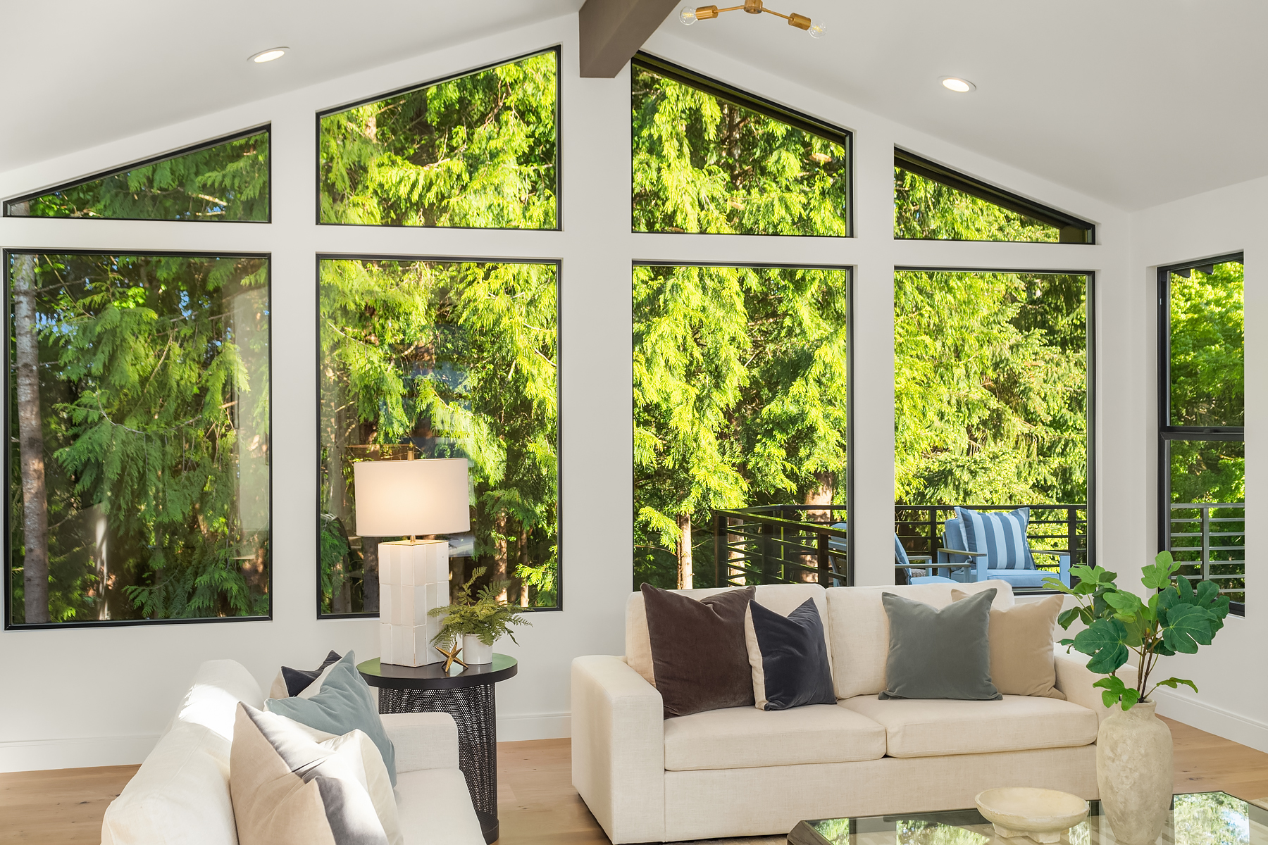 A bright living room with large, angled windows showing lush green trees outside. Two cream sofas with assorted pillows, a round black side table with a lamp, and decor fill the sunlit cozy space.