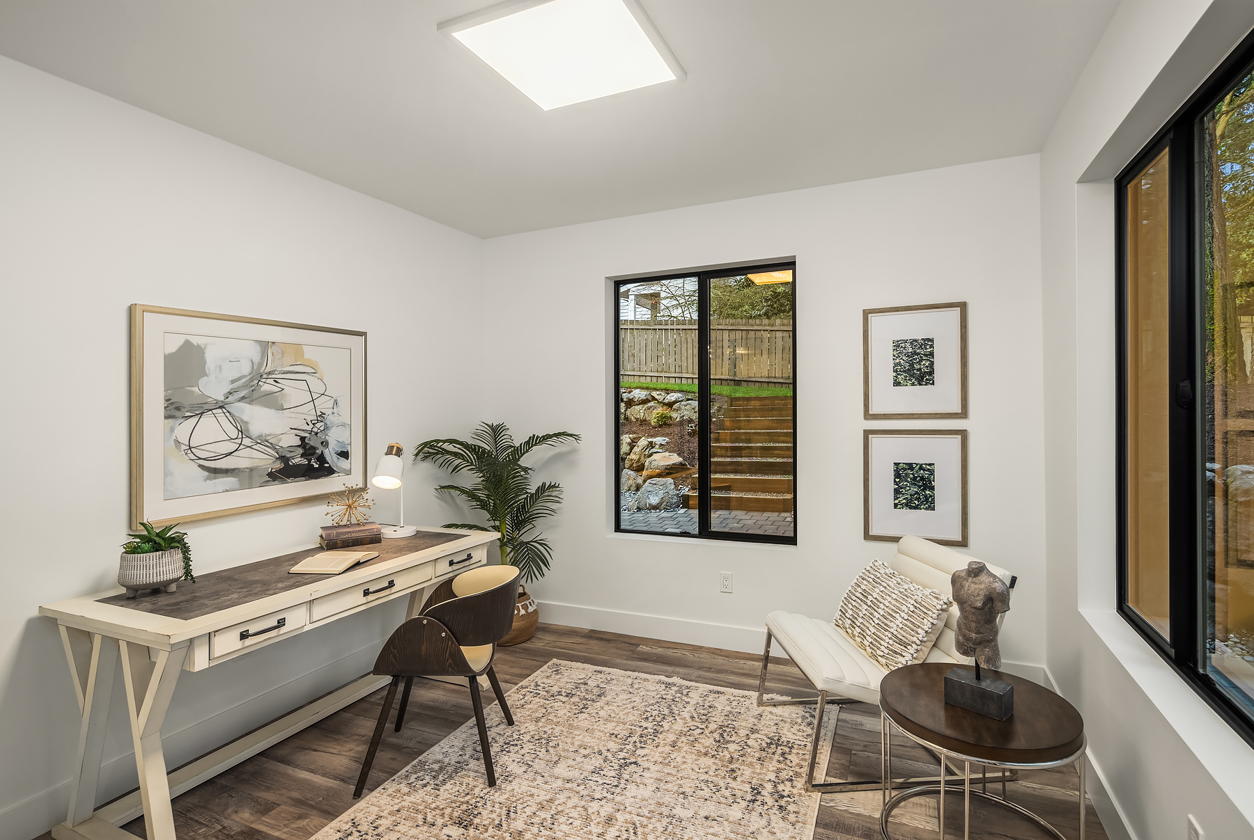 A modern home office with a wooden desk, chair, and lamp on the left, a plant in the corner, a rug, and a white lounge chair with a small table on the right. Large windows show a green outdoor space.