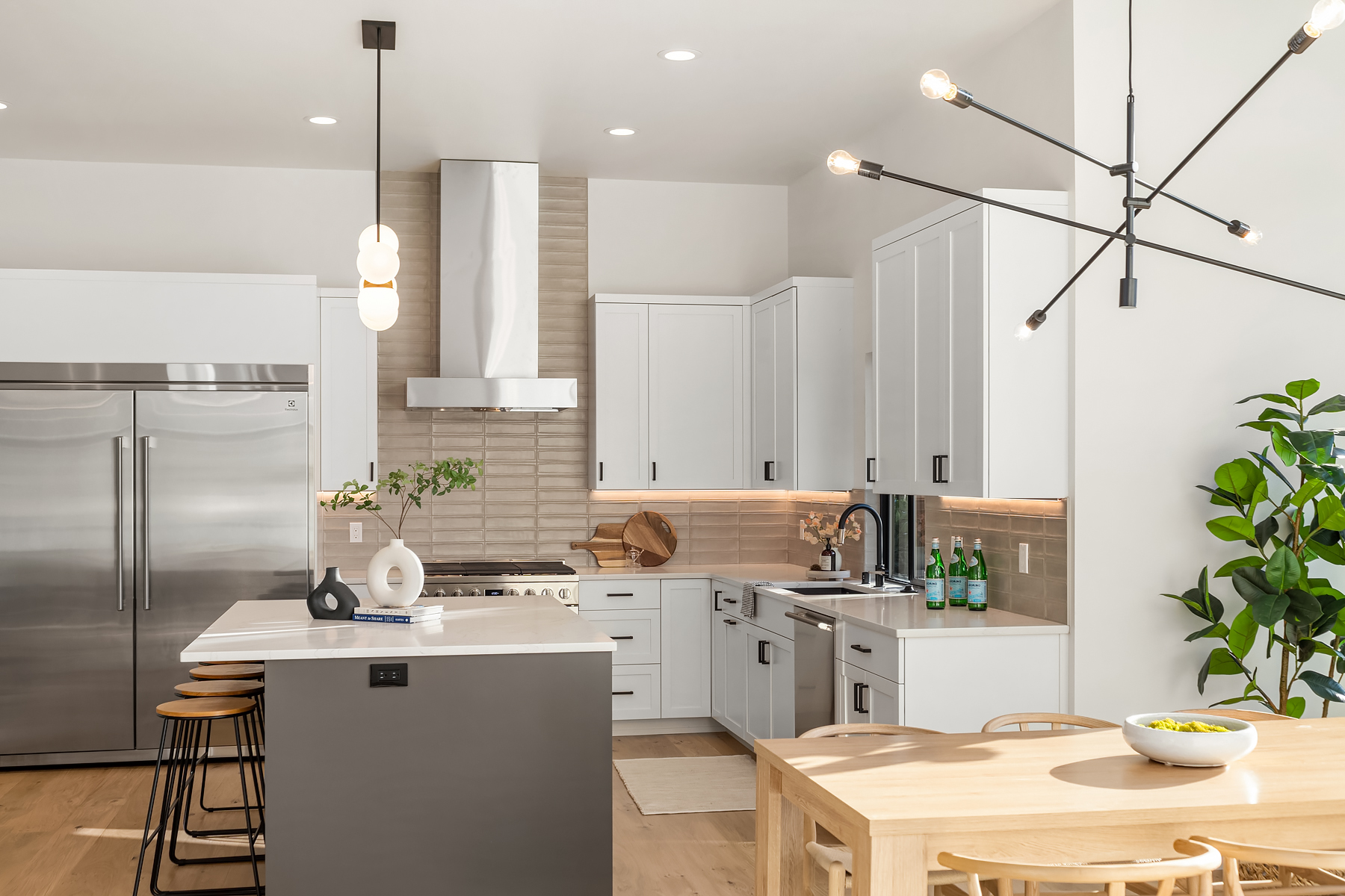 Modern kitchen with white cabinets, stainless steel appliances, a center island with barstools, and a wooden dining table. Pendant lights and a large plant add decorative touches. The space is bright with natural and artificial light.