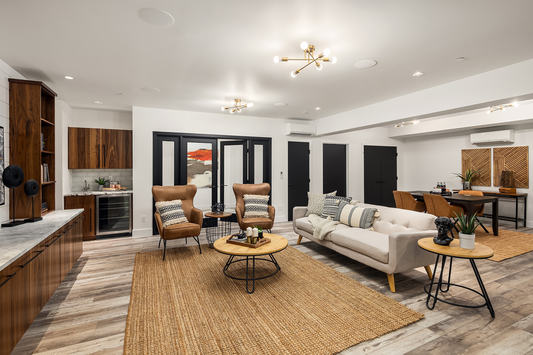 Modern living room with neutral tones, featuring a white sofa, two brown armchairs, round wooden tables, woven rugs, a dining area in the back, and wood cabinetry on the left. Black double doors and contemporary lighting complete the space.