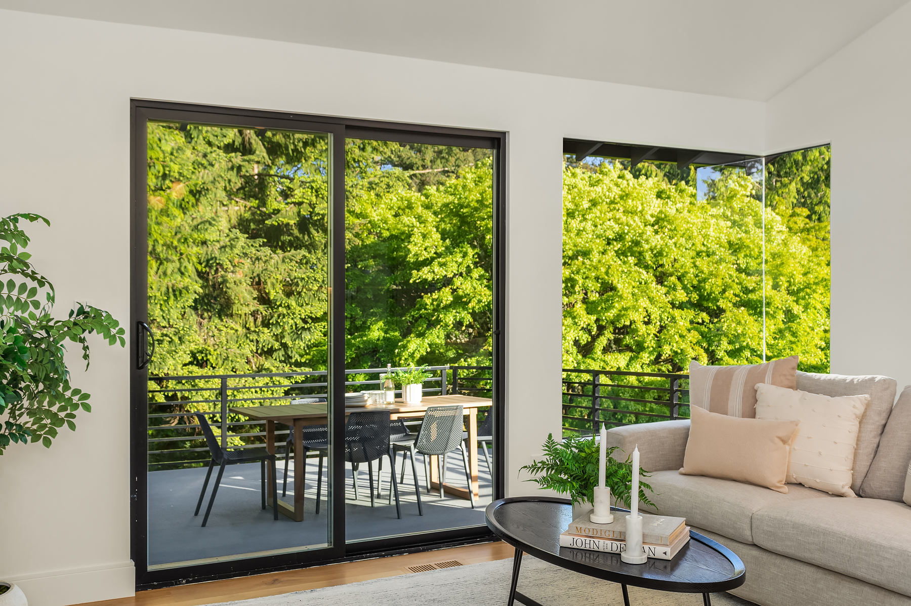 A bright living room with a beige sofa, a round coffee table, and large glass doors opening to a balcony with a dining set. Lush green trees are visible outside, filling the space with natural light.