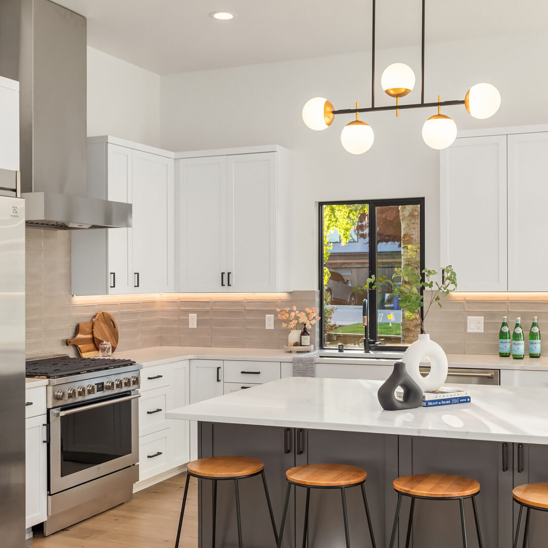 Modern kitchen with white cabinets, stainless steel appliances, a large island with three wooden stools, a geometric light fixture, and decorative items on the counters and island. A window lets in natural light.