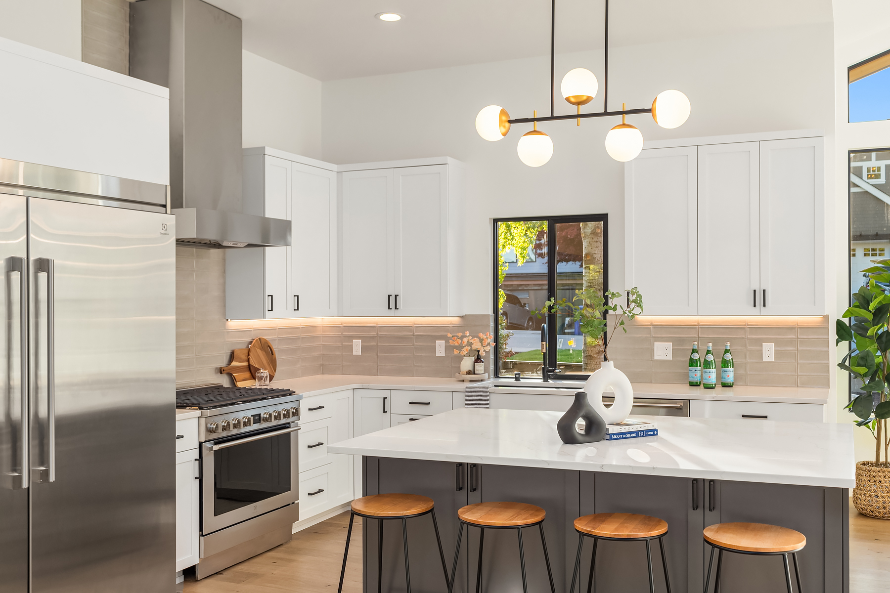 Modern kitchen with white cabinets, stainless steel appliances, a large island with three wooden stools, a geometric light fixture, and decorative items on the counters and island. A window lets in natural light.