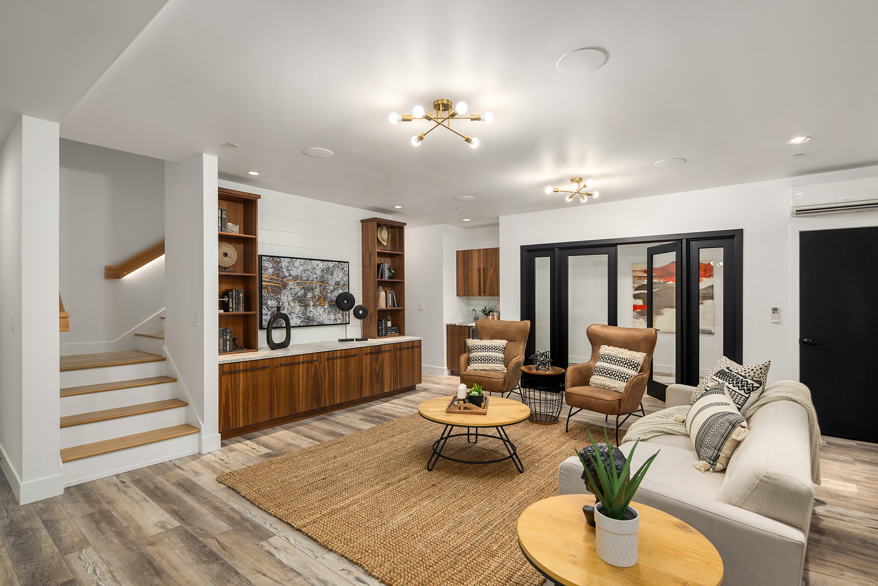 Modern living room with wood flooring, beige sofa, two brown armchairs, a large rug, wall-mounted TV, built-in wood cabinets, decorative items, and staircase; bright lighting and neutral color palette.