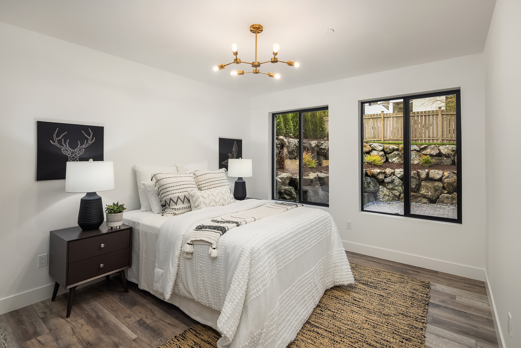 A modern bedroom with a neatly made bed, two bedside tables with lamps, abstract deer artwork, a geometric light fixture, large windows, and a view of a rocky garden outside. The decor is minimalist in neutral tones.