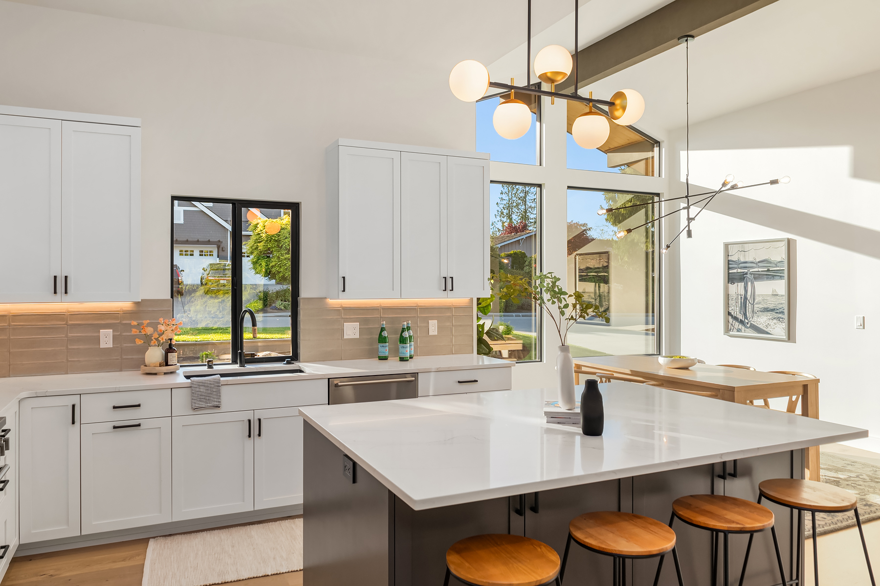 Bright, modern kitchen with white cabinets, a large island with wooden stools, pendant lighting, and a dining area by large windows letting in natural light. There are bottles and minimal decor on the countertops.