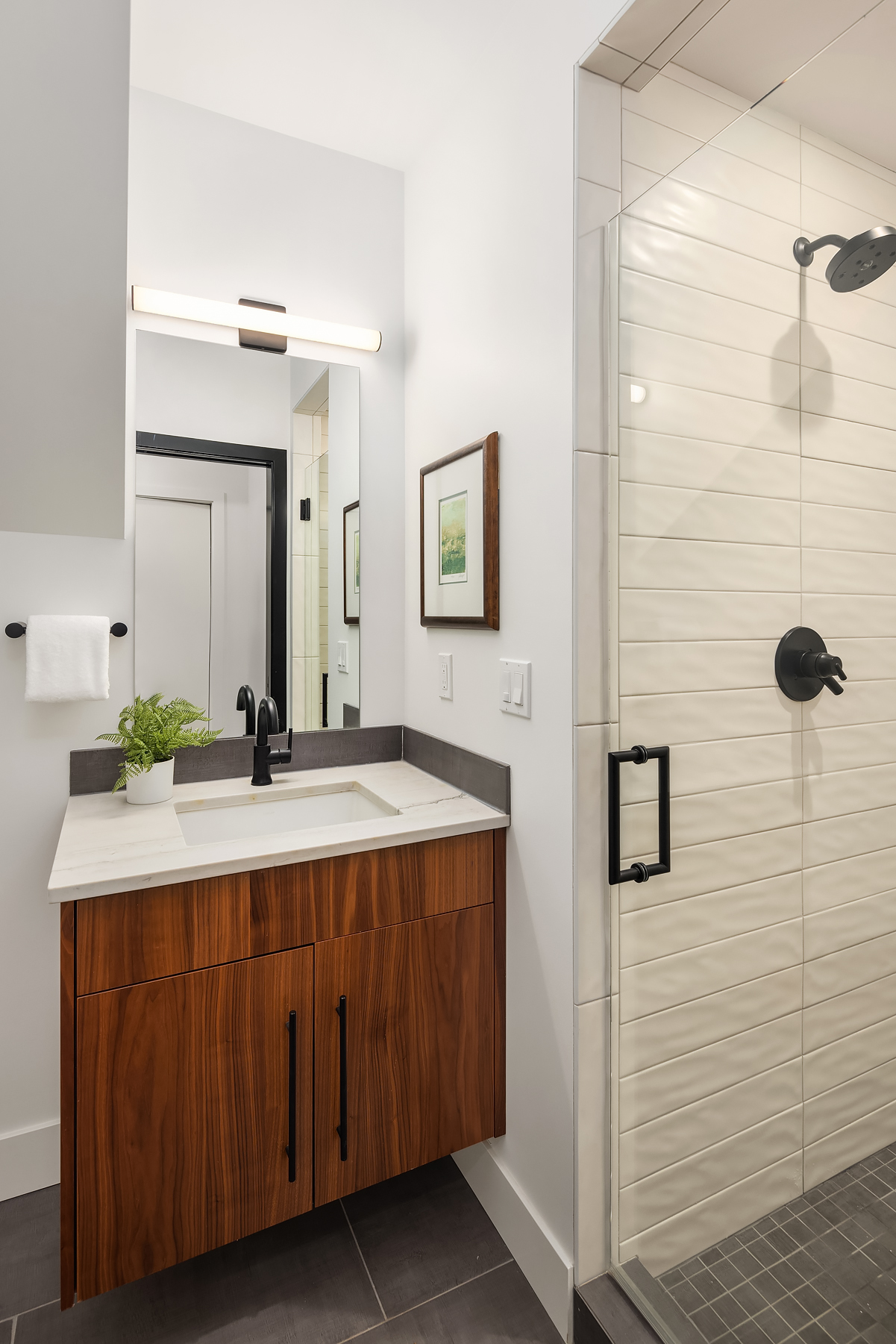 A modern bathroom with a wooden vanity, white countertop, black fixtures, a rectangular mirror, a potted plant, a towel ring, framed artwork, and a glass shower with white subway tile and black hardware.