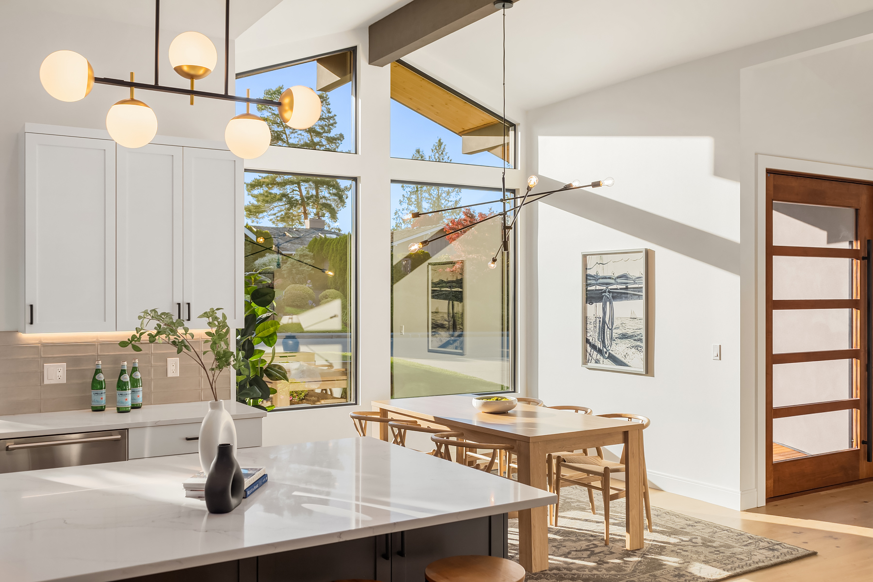 Bright, modern kitchen and dining area with large windows, light wood furniture, white cabinets, marble countertops, and contemporary pendant lighting. Sunlight streams in, highlighting a minimalist, open design.