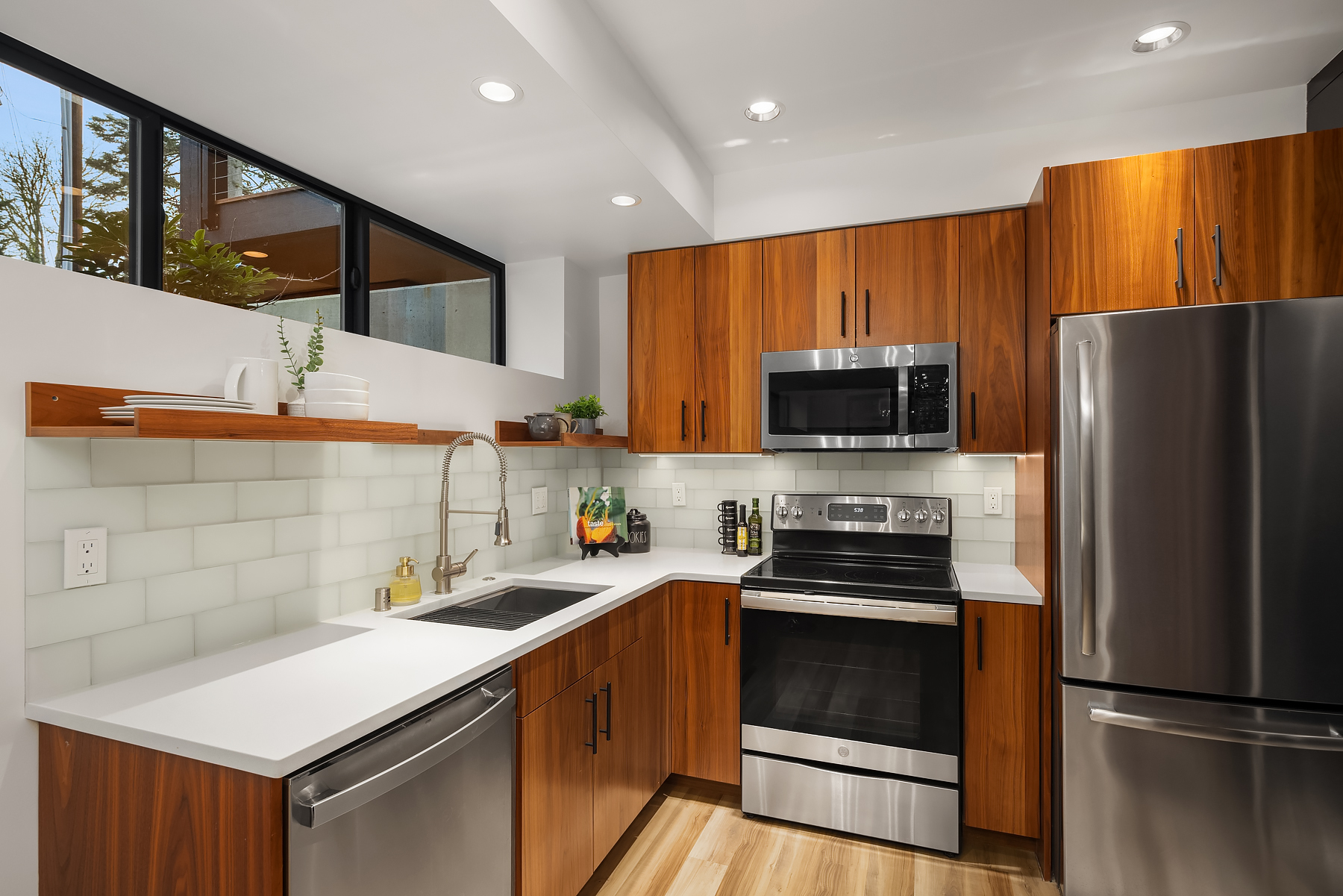 Modern kitchen with stainless steel appliances, wooden cabinets, white countertops, a tiled backsplash, and open shelves with plants and decor. Large window brings in natural light.
