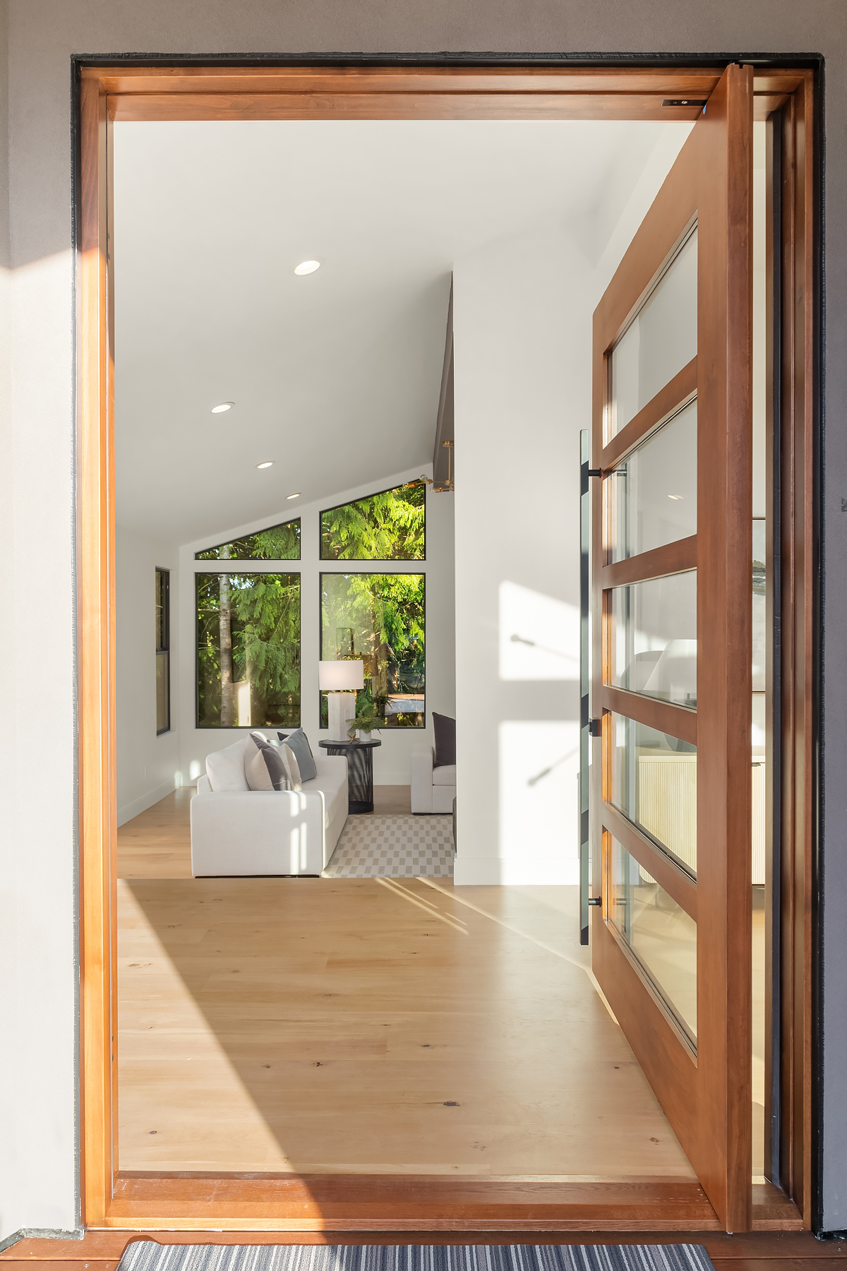 Open wooden door leading into a modern, bright living room with large windows, light wood flooring, a white sofa, and green trees visible outside. Natural light fills the spacious, minimalist interior.