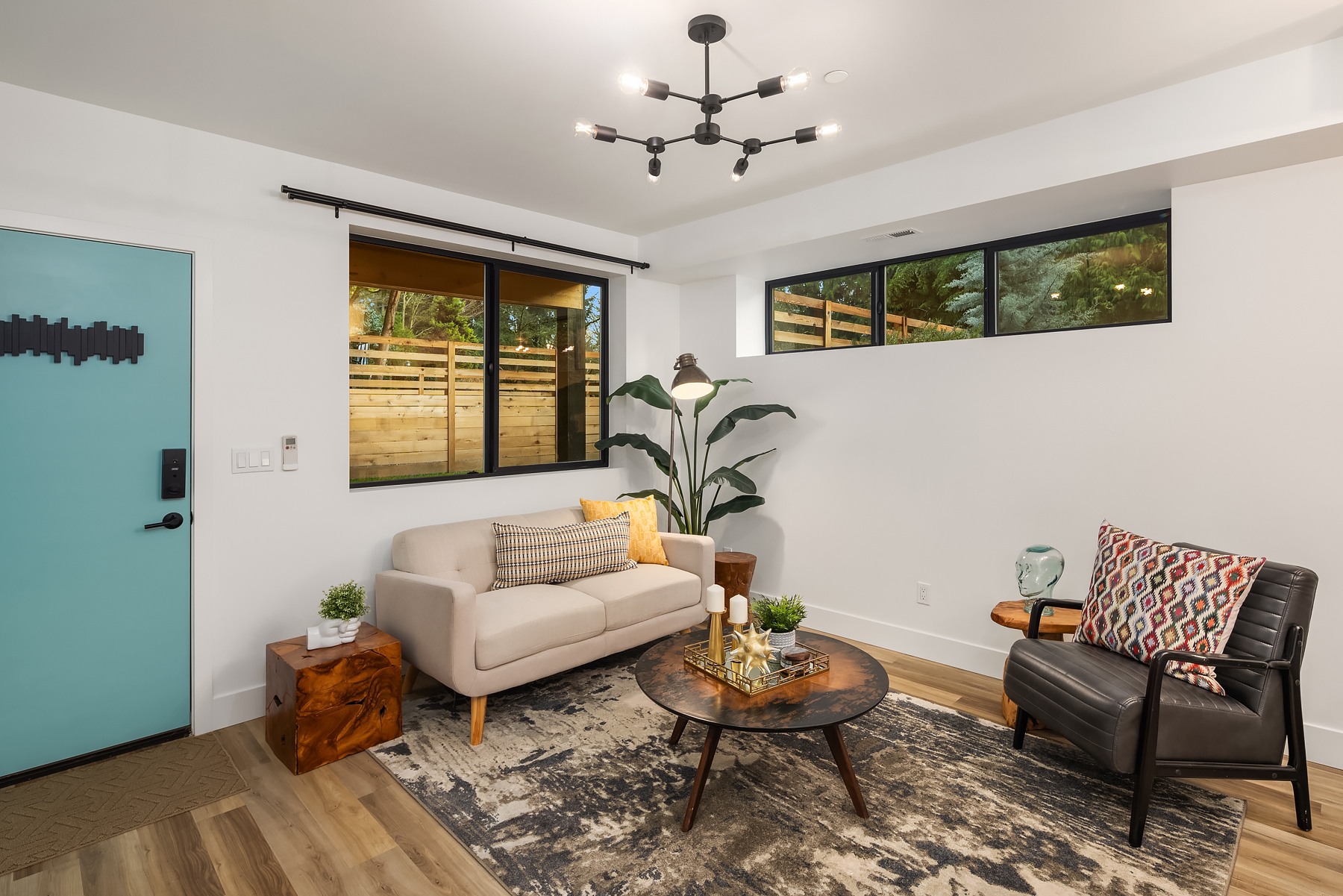 Modern living room with a light beige sofa, black armchair, round coffee table, large plant, and floor lamp. Blue front door and large window show a wooden fence and greenery outside. Warm, stylish decor on wood floors.