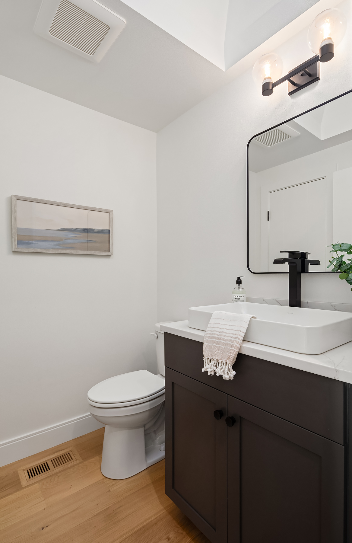 A modern bathroom with a white toilet, black vanity with a white rectangular sink, black faucet, hand towel, soap dispenser, square mirror, light fixture, artwork on the wall, and light wood floor.