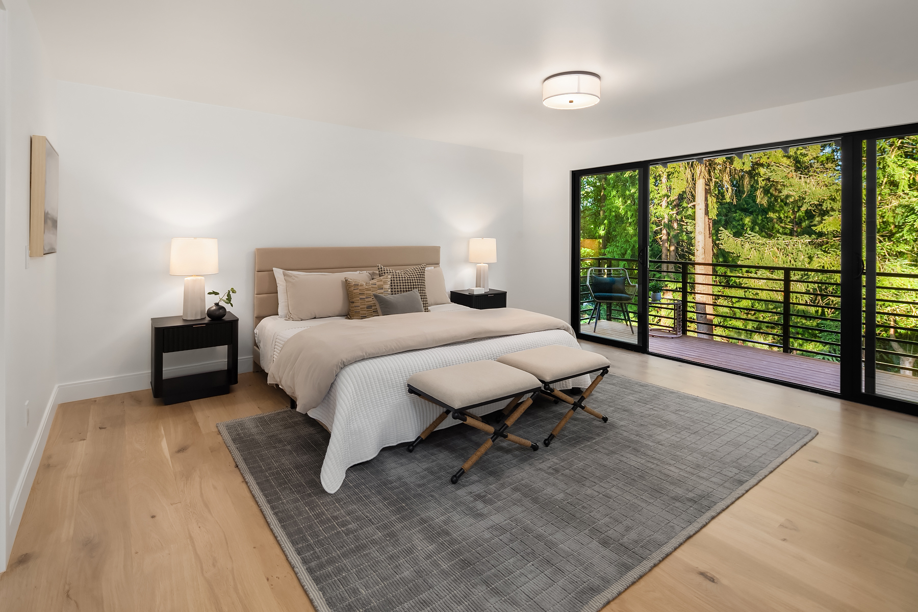 A modern bedroom with a large bed, two benches at the foot, two bedside tables with lamps, a gray rug, wood flooring, and glass doors opening to a balcony with a view of trees.