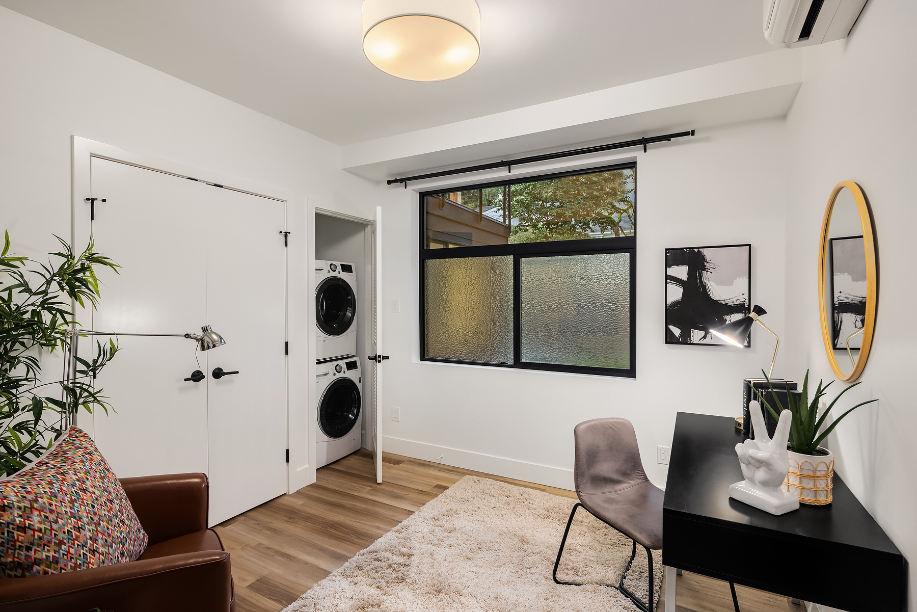 Modern home office with a black desk, chair, and decor, a brown armchair with a colorful pillow, indoor plants, a window with frosted glass, and a closet with stacked washer and dryer units.
