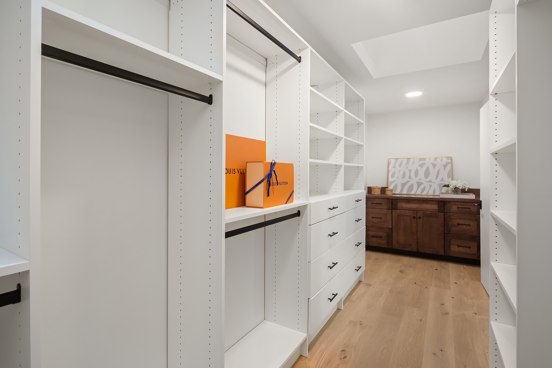 A modern walk-in closet with white shelves and drawers, a wooden floor, a Louis Vuitton box on a shelf, and a wooden dresser with decor at the far end under a skylight.
