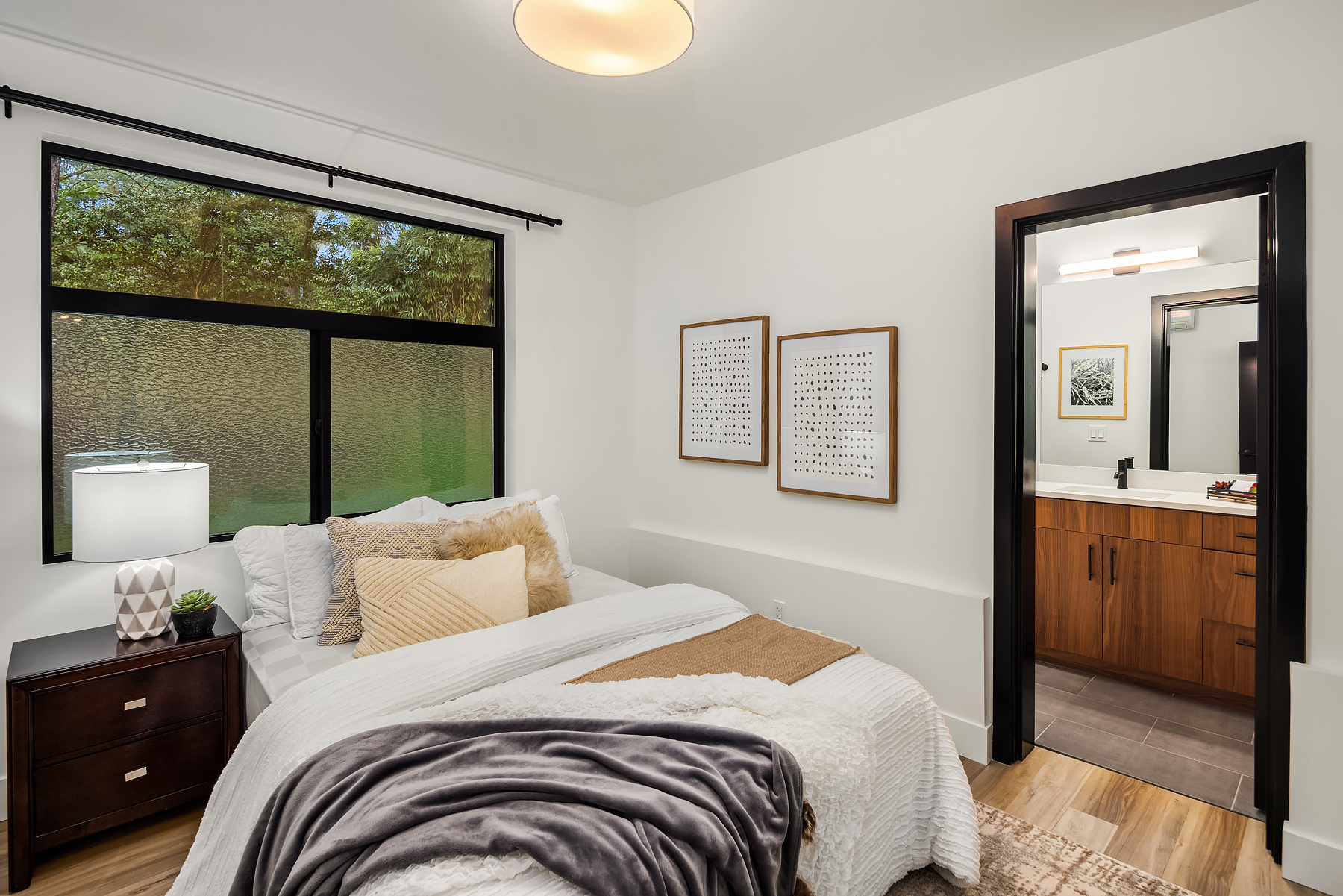 A modern, cozy bedroom with a neatly made bed, brown wooden nightstand, large window, and an open door leading to a bathroom with a wooden vanity and mirror. Two framed prints hang on the white wall.