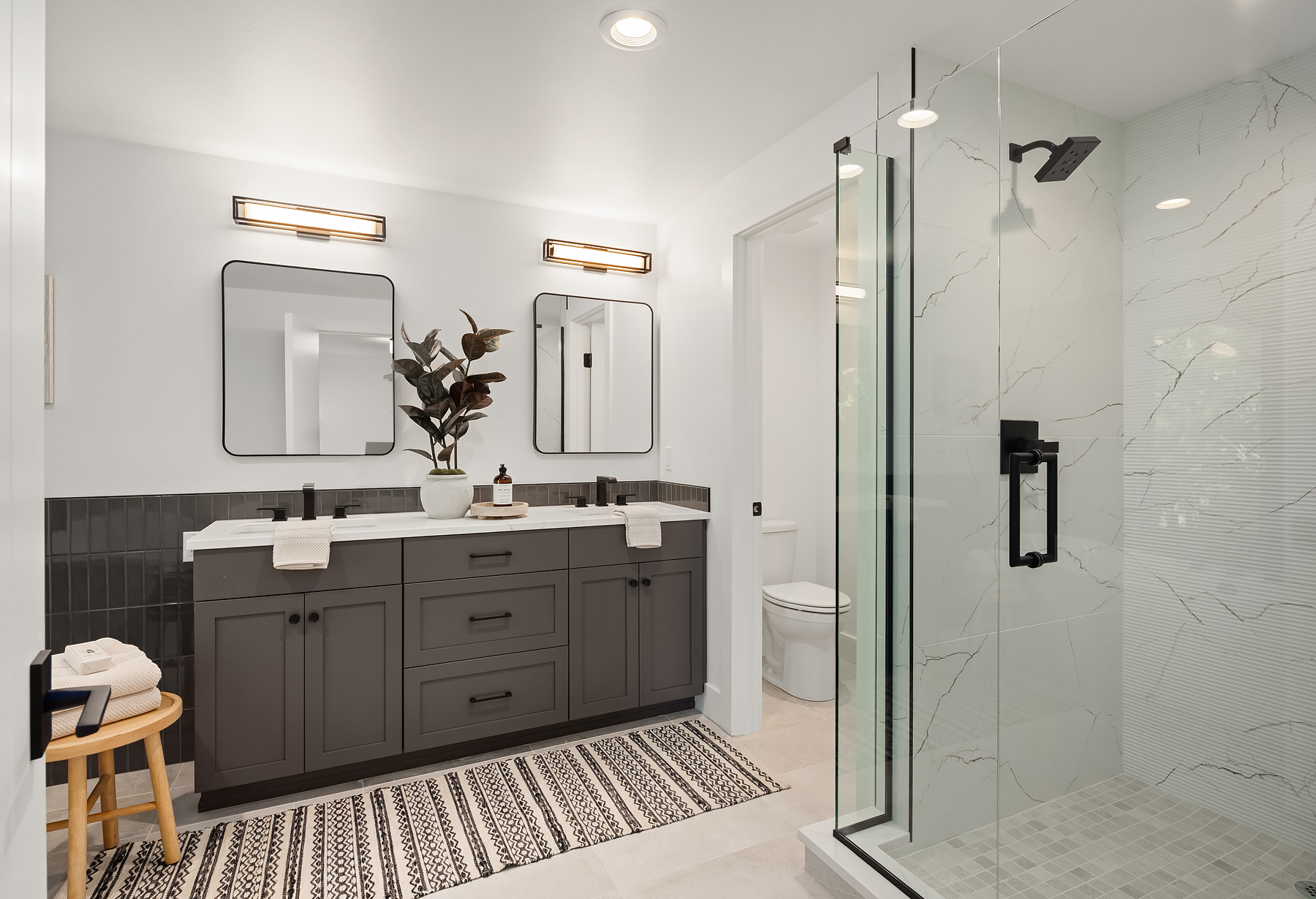 Modern bathroom with double-sink vanity, two mirrors, a plant, a glass-enclosed shower with black fixtures, a toilet, and a wooden stool with folded towels on a patterned rug.