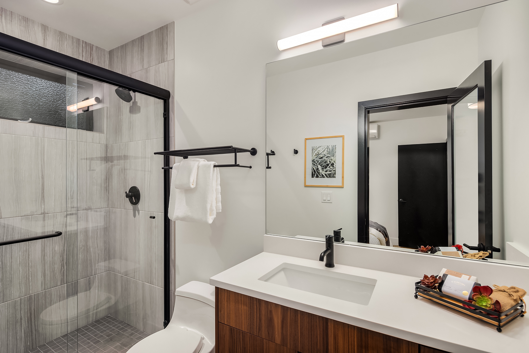Modern bathroom with a glass-enclosed shower, black fixtures, white countertop with a sink, wooden vanity, mirror, wall-mounted light, towel rack with white towels, and a tray with toiletries and fruit on the counter.