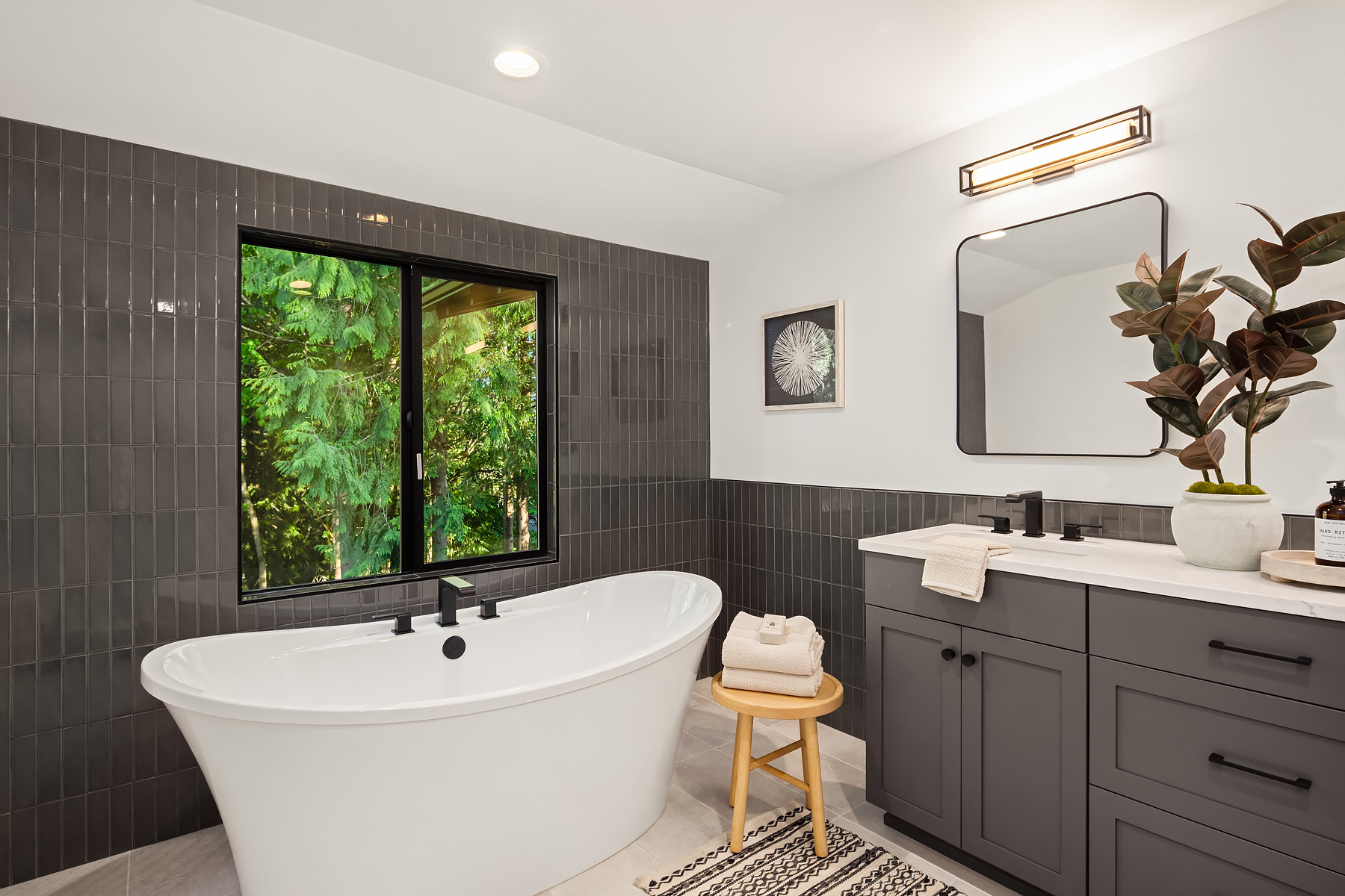 Modern bathroom with a white freestanding tub, dark tile accent wall, large window showing greenery, gray vanity with a square mirror, potted plant, and folded towels on a wooden stool.