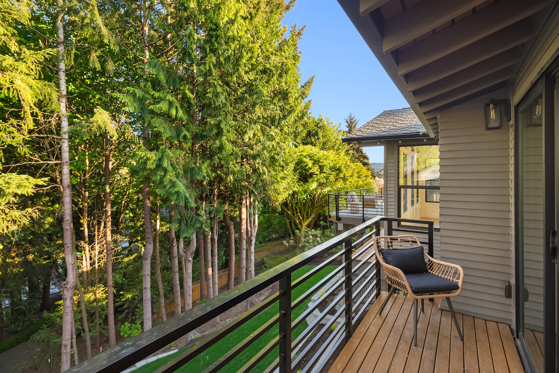 A wooden balcony with a black metal railing overlooks a green, tree-filled yard. The balcony has a single wicker chair with a cushion, and the house exterior is beige with siding. The scene is bright and sunny.