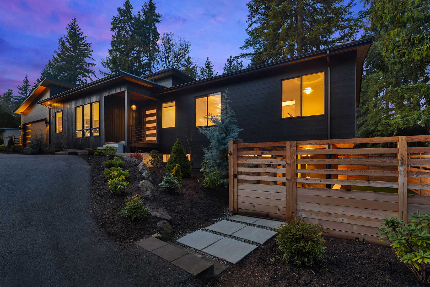 Modern single-story house with dark wood siding, large lit windows, and landscaped yard, surrounded by tall trees at dusk. A wooden gate and paved walkway lead to the entrance, with warm lights glowing inside.