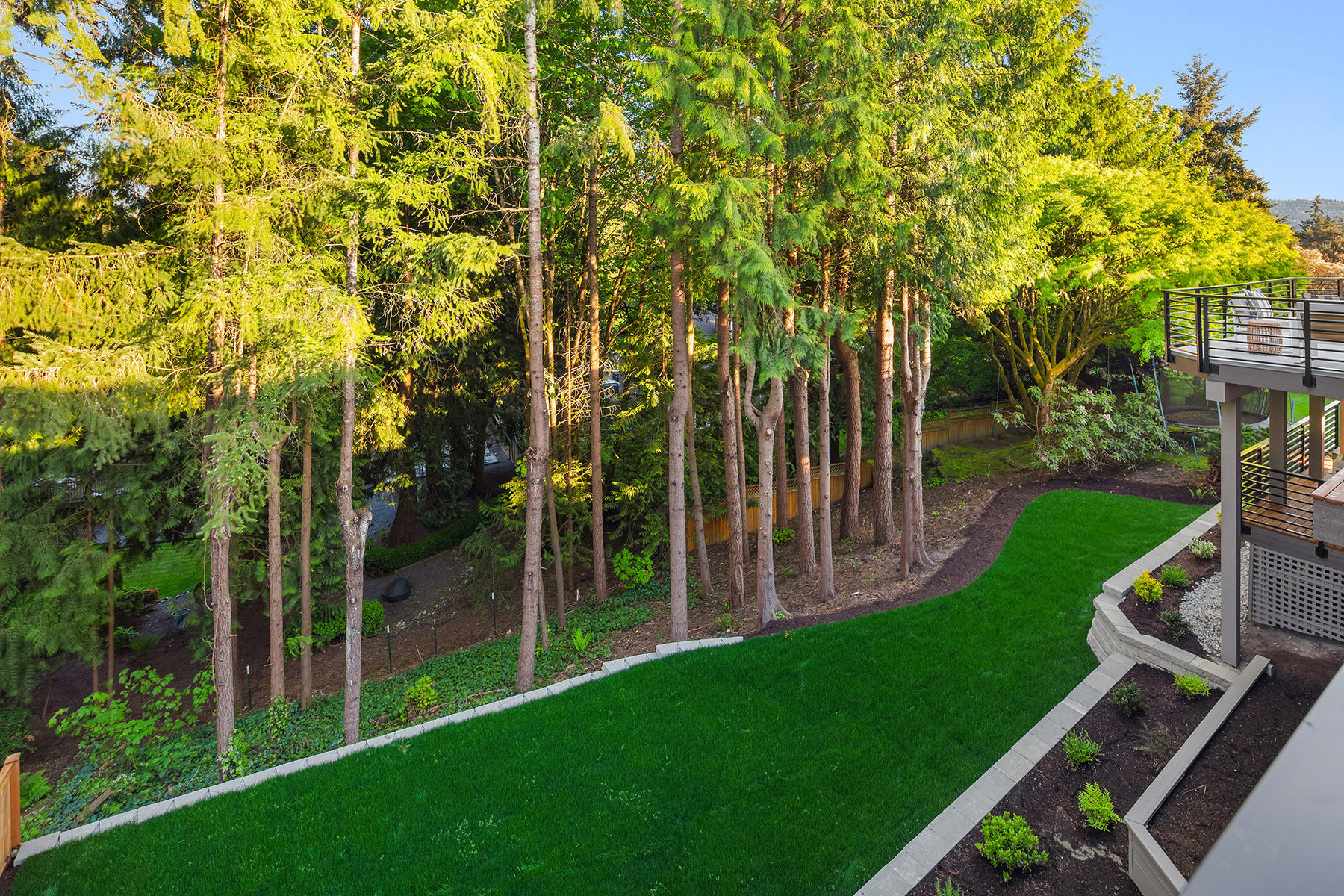A backyard featuring a lush green lawn bordered by a landscaped garden and tall trees, with a deck and railing visible on the right side under bright daylight.