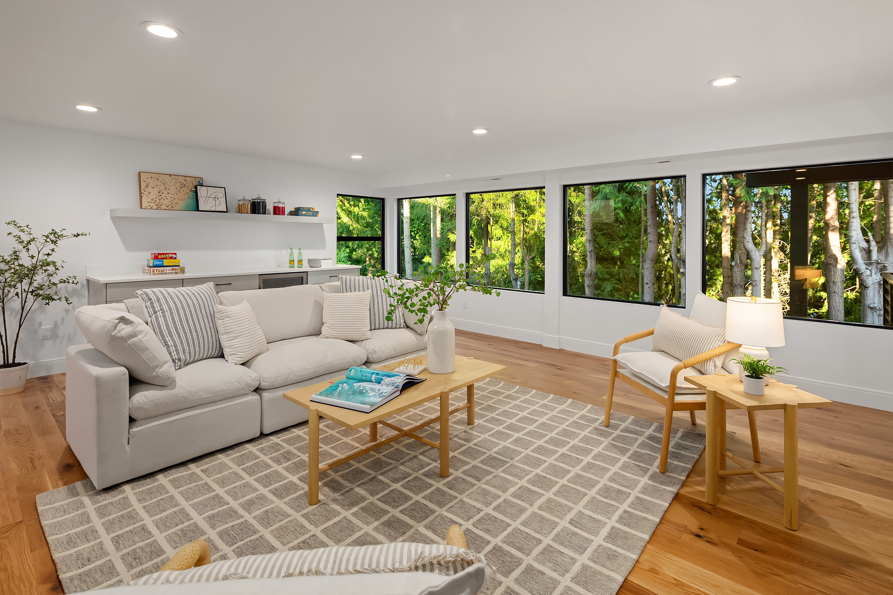 Bright, modern living room with light wood floors, white walls, a soft beige sofa, armchair, and a wooden coffee table. Large windows reveal lush green trees outside, letting in natural light. Decor includes plants and books.