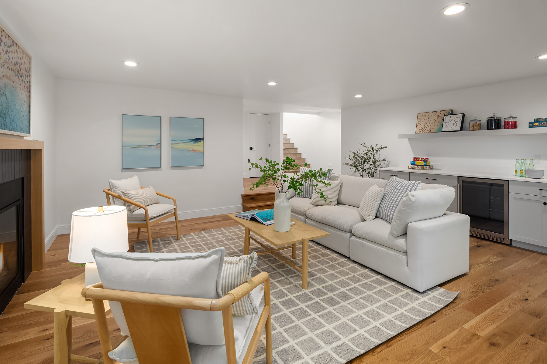 A modern living room with a white sofa, two wooden chairs, a coffee table, and a patterned rug. The space has hardwood floors, wall art, built-in shelving, and bright, recessed lighting. A staircase leads up from the back.