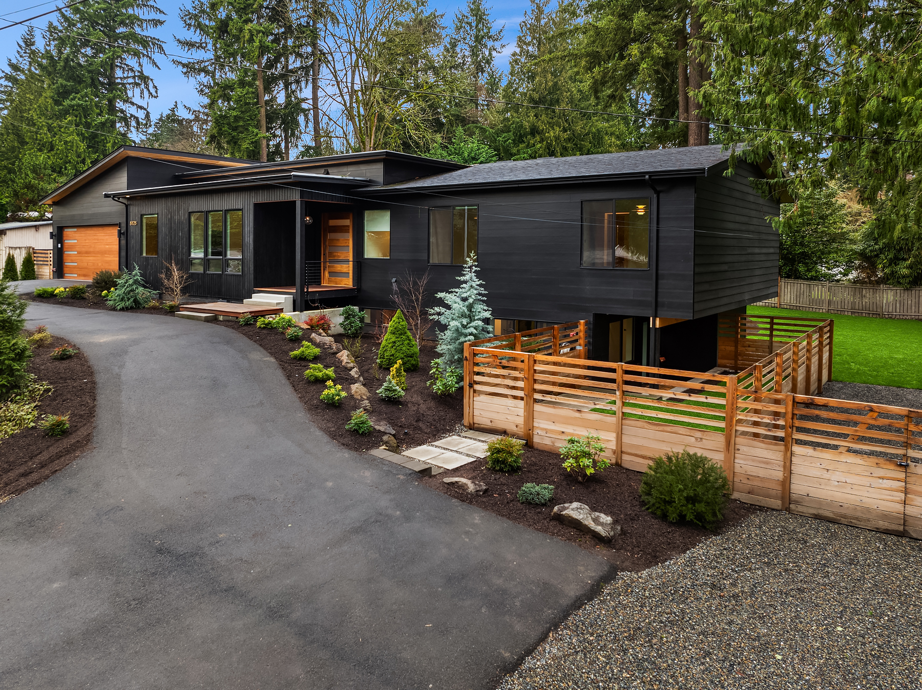 Modern black house with large windows, surrounded by lush landscaping, trees, and a wooden fence. A paved driveway leads to a garage, and a well-kept lawn is visible to the side.