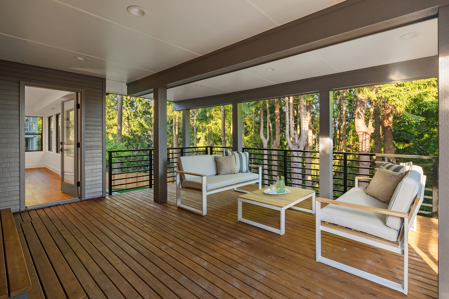 Covered outdoor deck with wooden floor, two modern white sofas, a coffee table with drinks, and a bench. Surrounded by trees and greenery, the space looks inviting and peaceful with natural light streaming in.