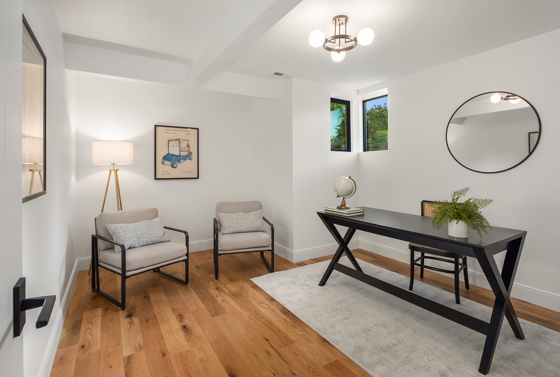 Modern home office with a black desk, globe, plant, and books on a gray rug, two beige armchairs, framed artwork, a floor lamp, and a round mirror on white walls with wood floors and two small windows.