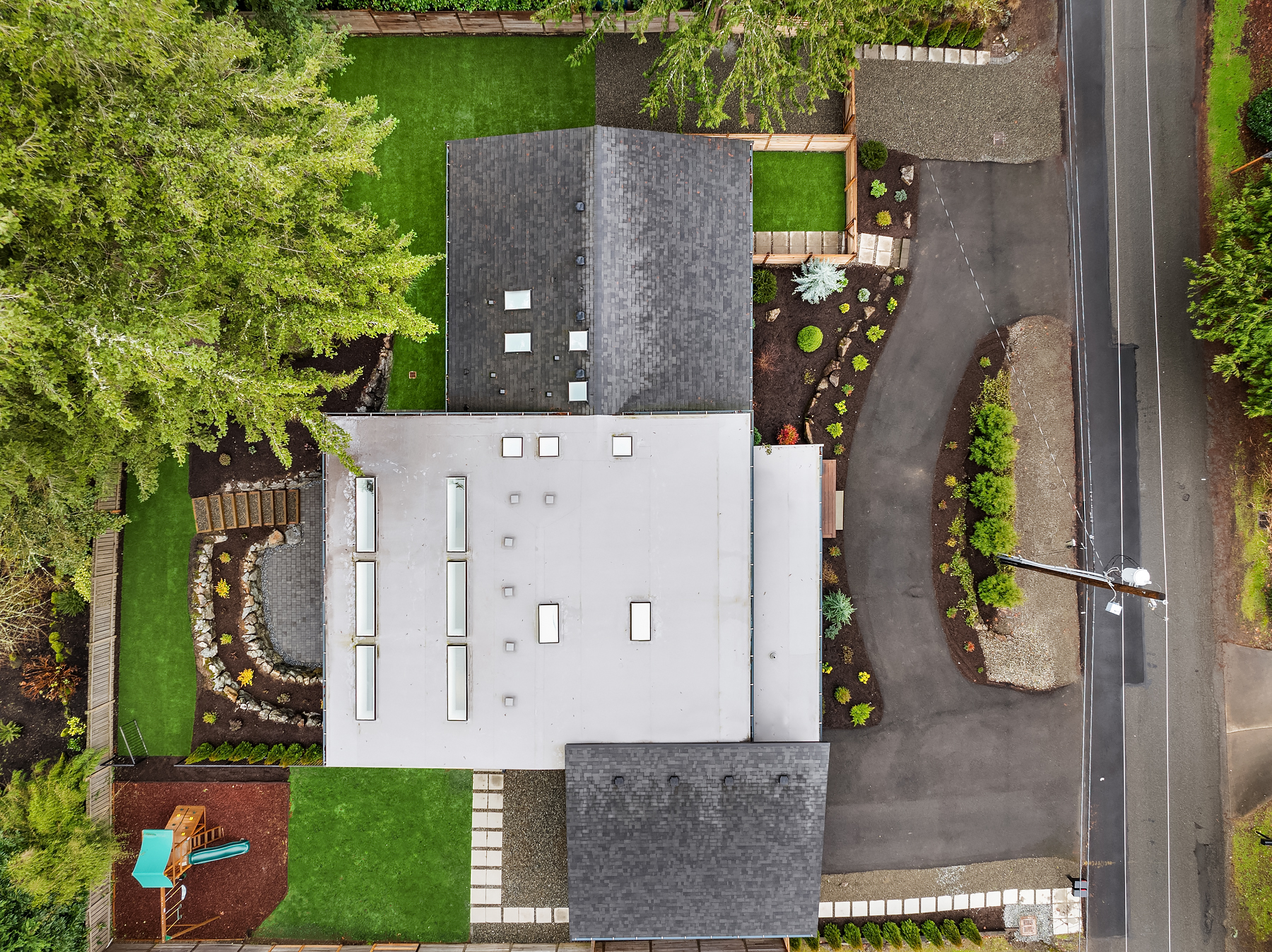 Aerial view of a house with a gray roof surrounded by a landscaped yard, a curved driveway, green lawns, trees, and a playground set in the lower left corner. The property is bordered by fences and streets.