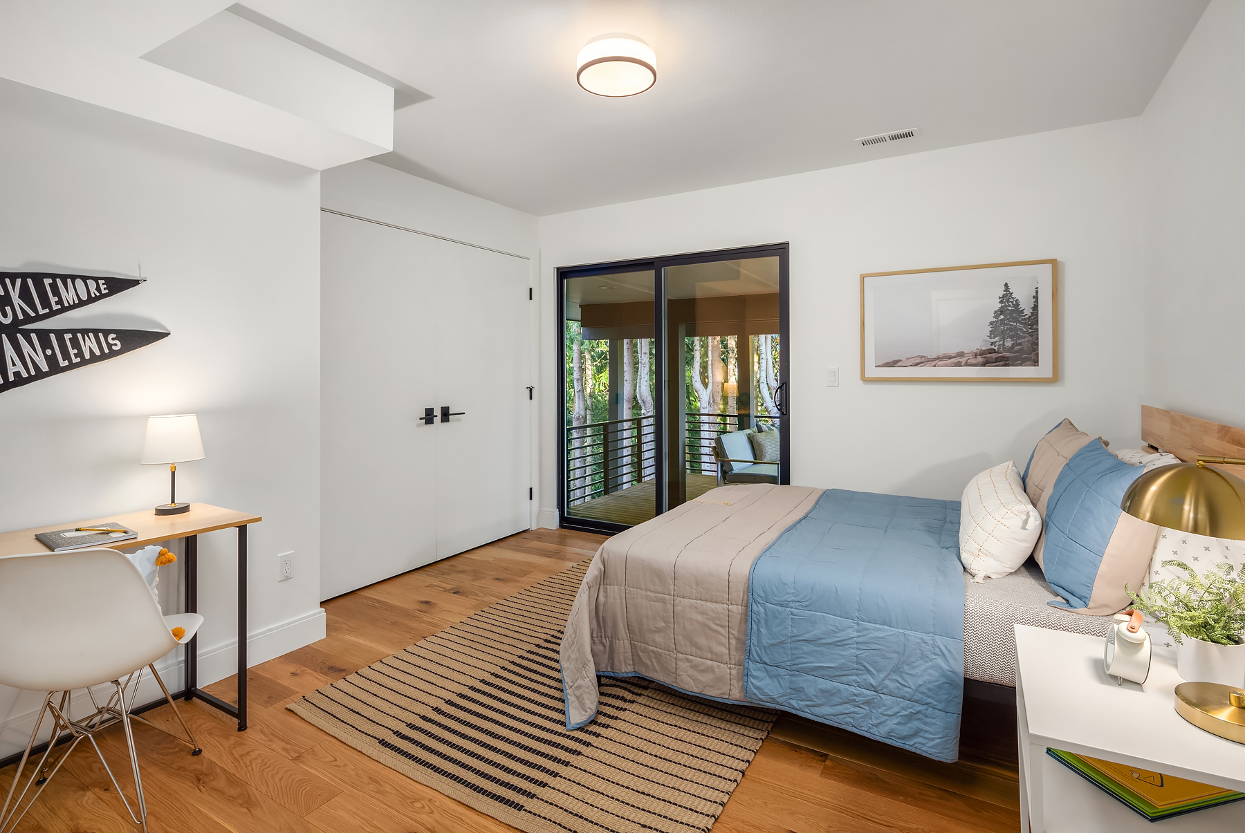 A modern bedroom with a bed covered in blue and beige bedding, a striped rug, a white desk with a lamp and chair, and a nightstand with a gold lamp. Glass doors open to a balcony with trees outside.