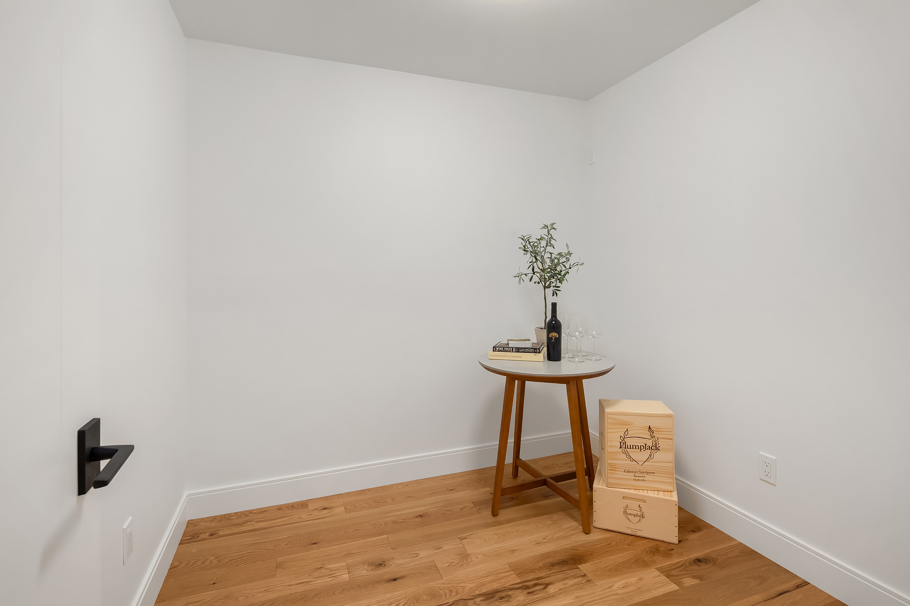 A small, empty room with white walls and wooden floor, featuring a round table holding a wine bottle, glasses, books, and a plant. Two wooden crates are stacked beside the table.