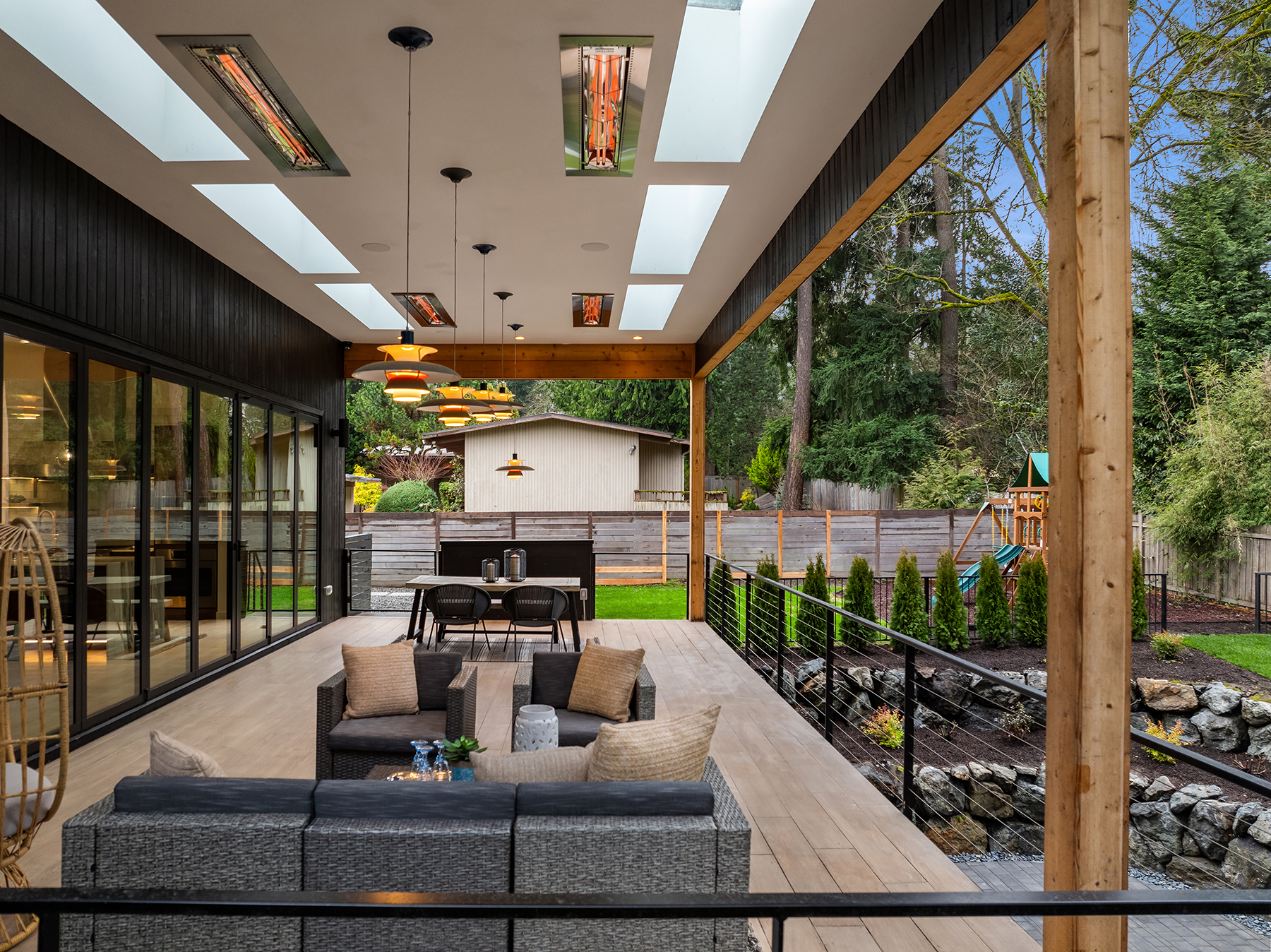 Modern covered patio with wicker seating, hanging lights, overhead heaters, and a dining table. The patio overlooks a landscaped backyard with a playset, greenery, and a wooden fence. Large glass doors connect to the indoor space.