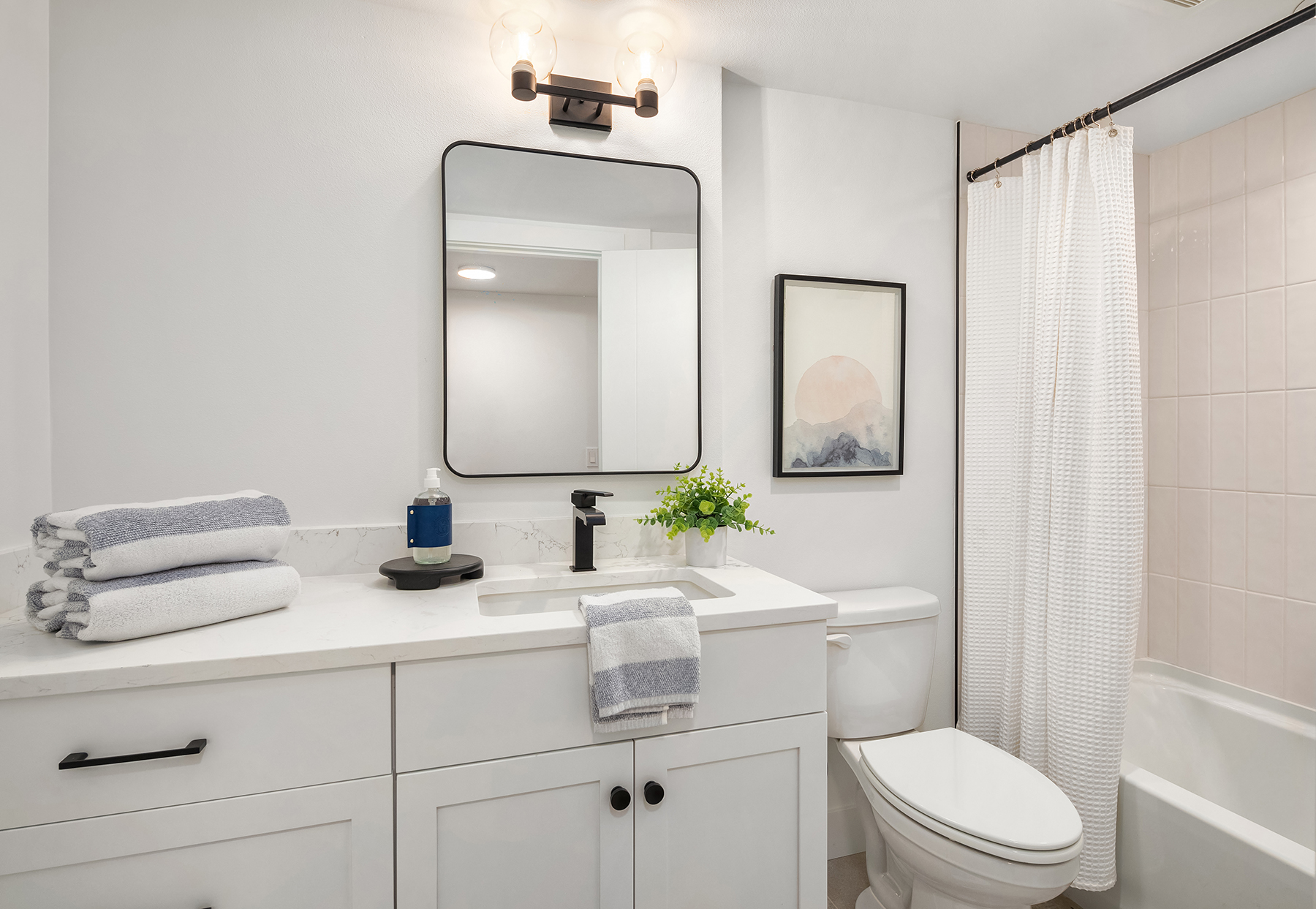 Modern bathroom with white cabinets, a black-framed mirror, stacked towels, a potted plant, and wall art. Bathtub with a white shower curtain is next to a white toilet under soft lighting.