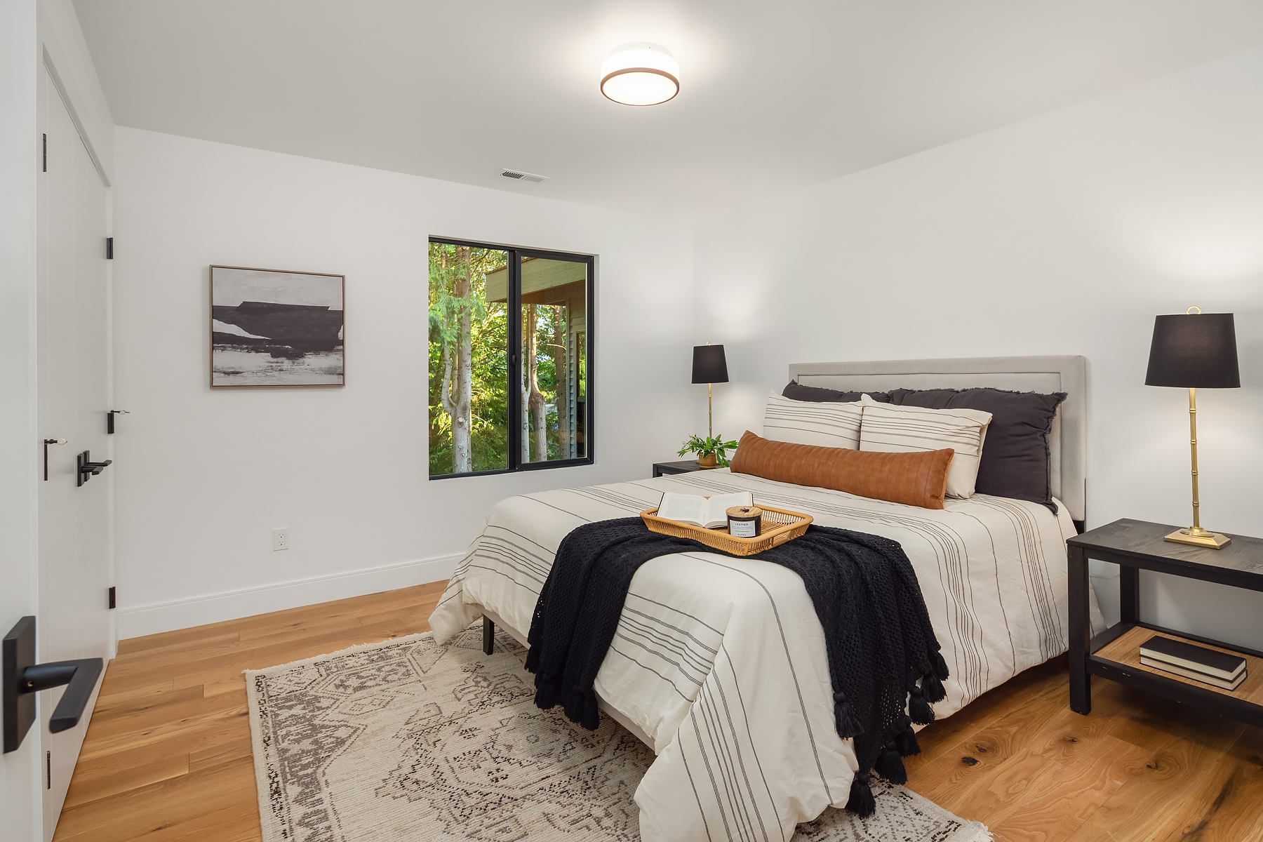 Modern bedroom with a neatly made bed, striped bedding, black throw, and decorative pillows. A tray is on the bed. The room has a rug, nightstands with lamps, a window, white walls, and wood flooring.