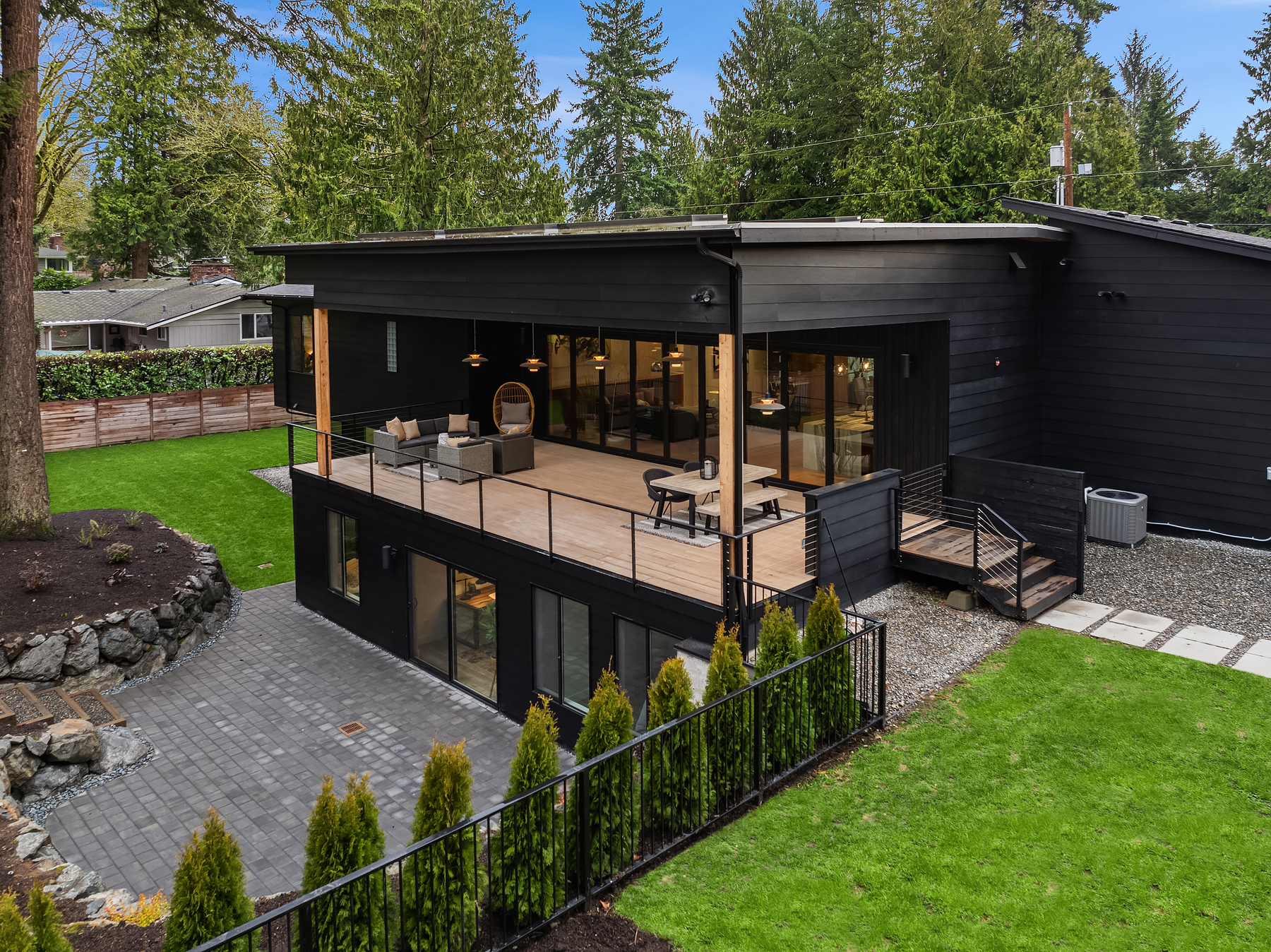 Modern black two-story house with large windows, a spacious covered deck, and patio below, surrounded by green lawn, trees, and landscaped garden beds in a suburban setting.