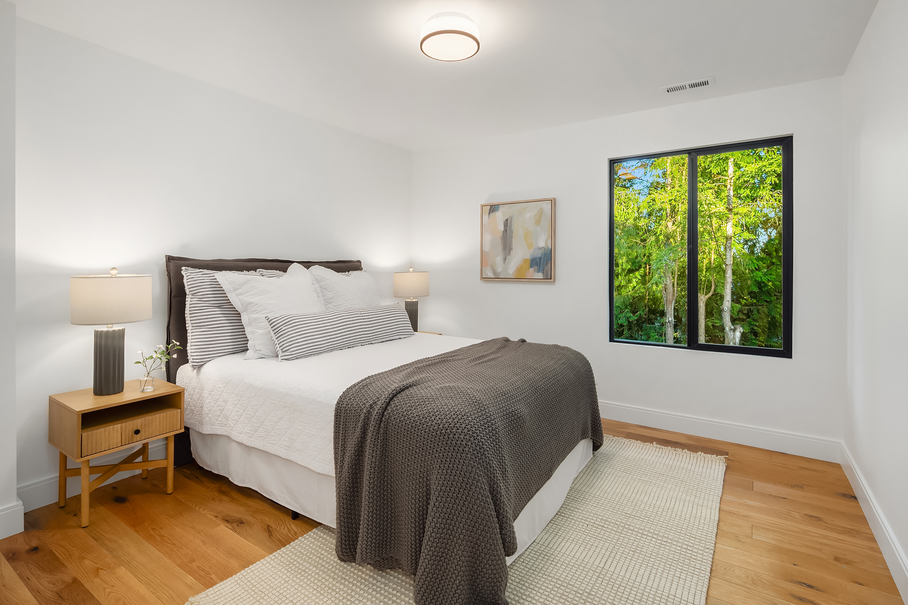 A bright, modern bedroom with a neatly made bed, gray blanket, striped pillows, wooden nightstand with a lamp and flower, abstract wall art, hardwood floors, and a large window showcasing green trees outside.