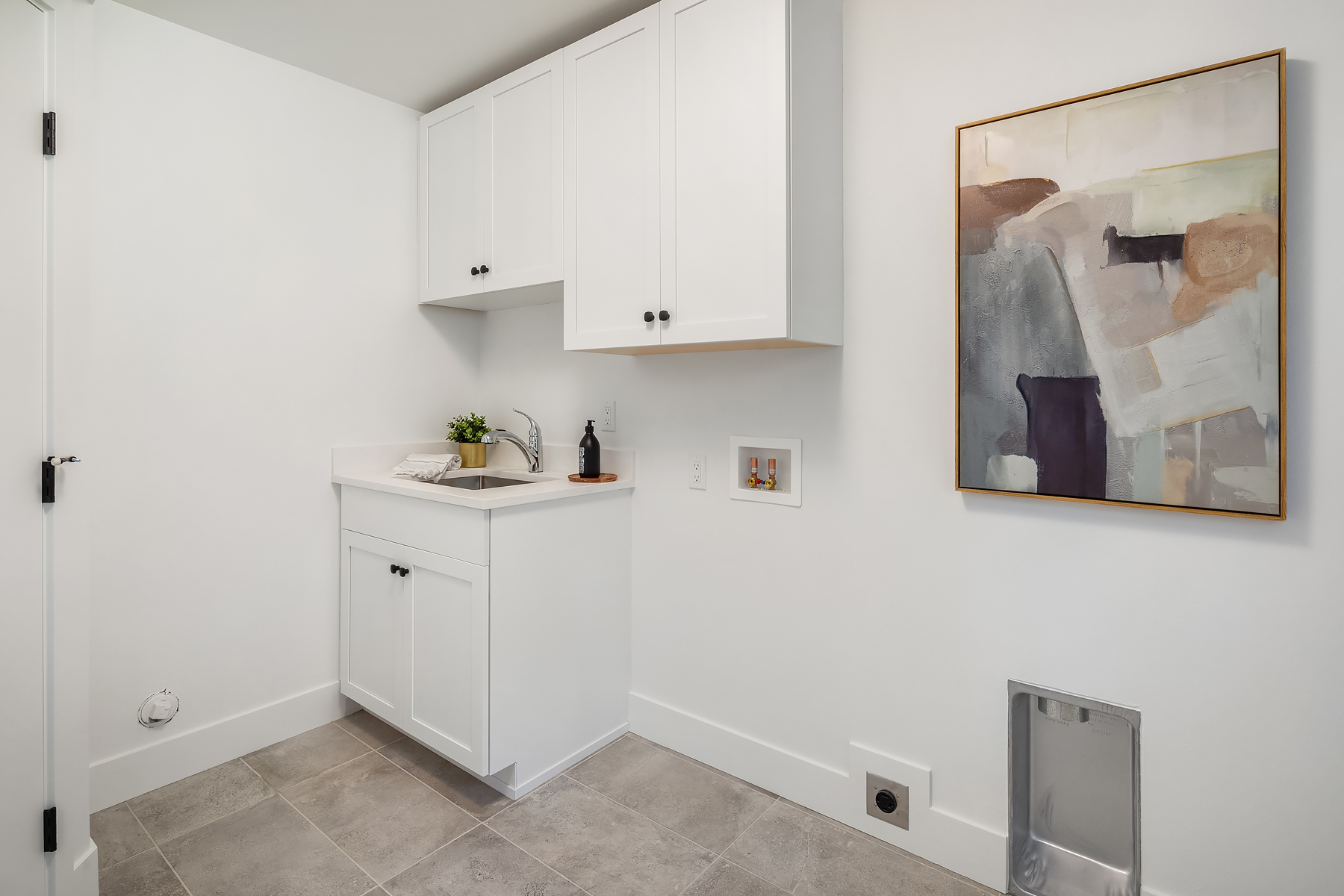 A modern laundry room with white cabinets, a countertop with a sink, a potted plant, and soap. Abstract art hangs on the white wall above gray tile flooring; washer and dryer hookups are visible.