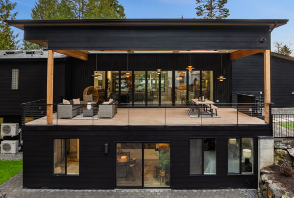 A modern black two-story house with a spacious upper deck featuring outdoor seating and dining areas, large glass doors, and wooden posts. The lower level has large windows and sits on a stone patio. Trees surround the home.