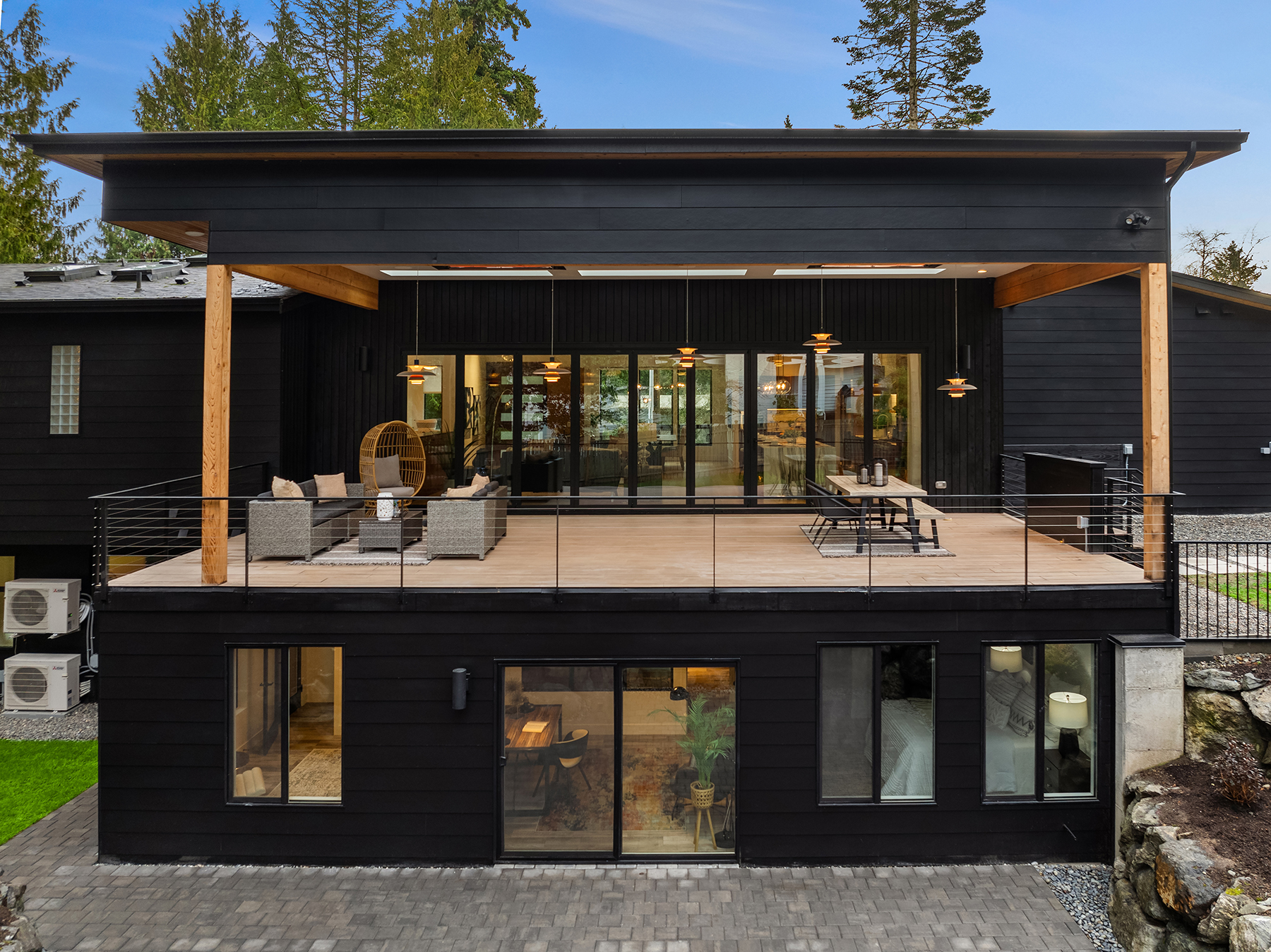 A modern black two-story house with a spacious upper deck featuring outdoor seating and dining areas, large glass doors, and wooden posts. The lower level has large windows and sits on a stone patio. Trees surround the home.
