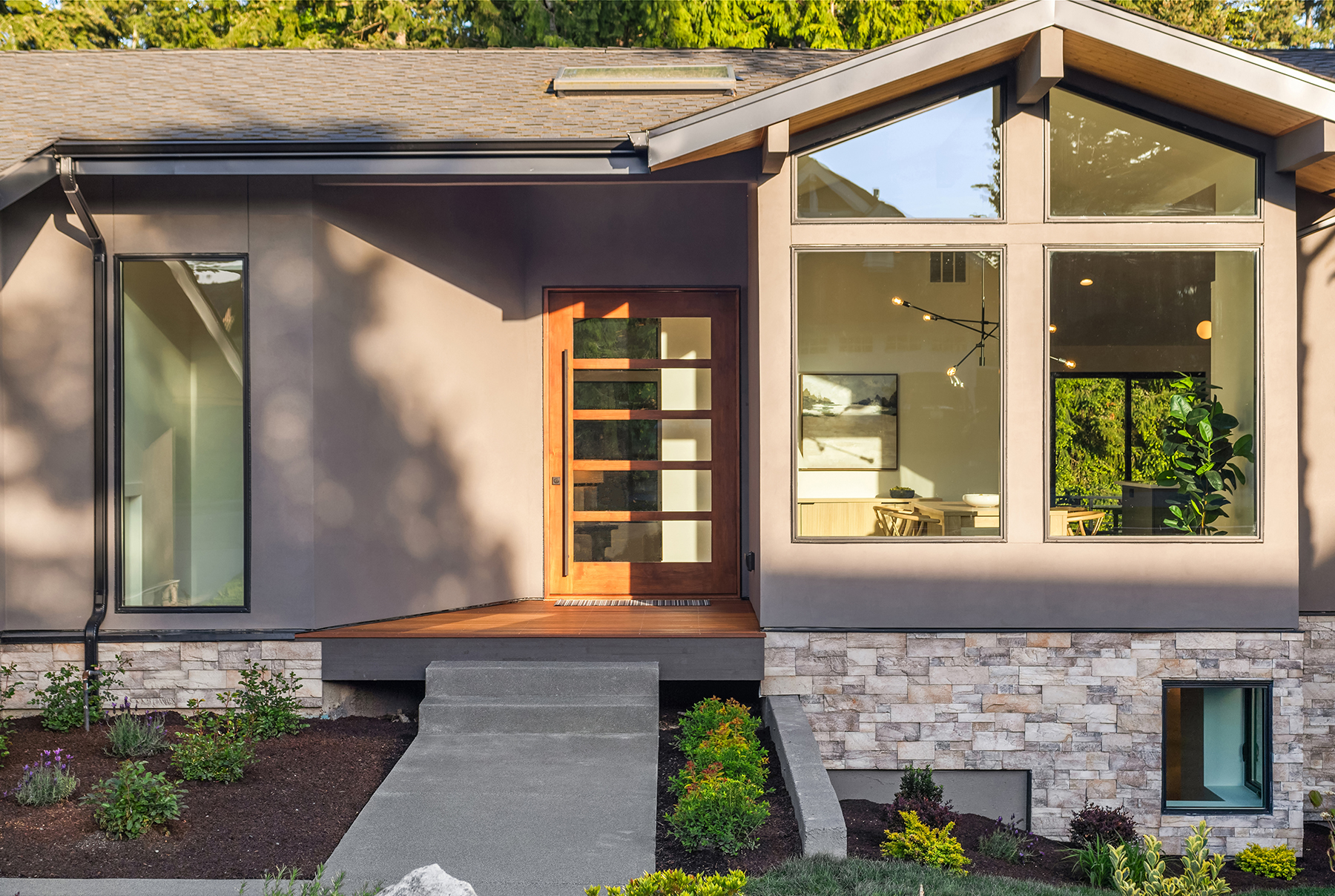 Modern house exterior with stone and stucco facade, large windows, wooden front door with glass panels, small porch, concrete walkway, and landscaped garden with shrubs and greenery.