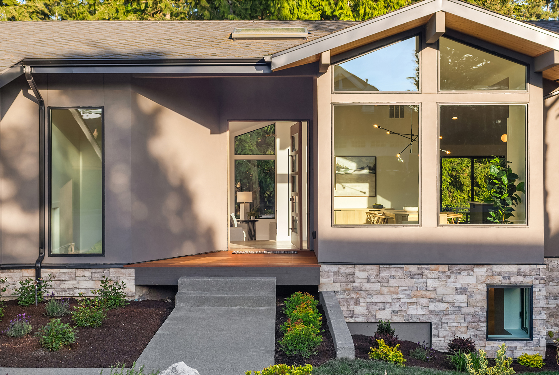 Modern house exterior with large windows, stone accents, a covered entryway, landscaped garden, and pathway leading to the front door. Warm light shines from inside, highlighting contemporary decor.