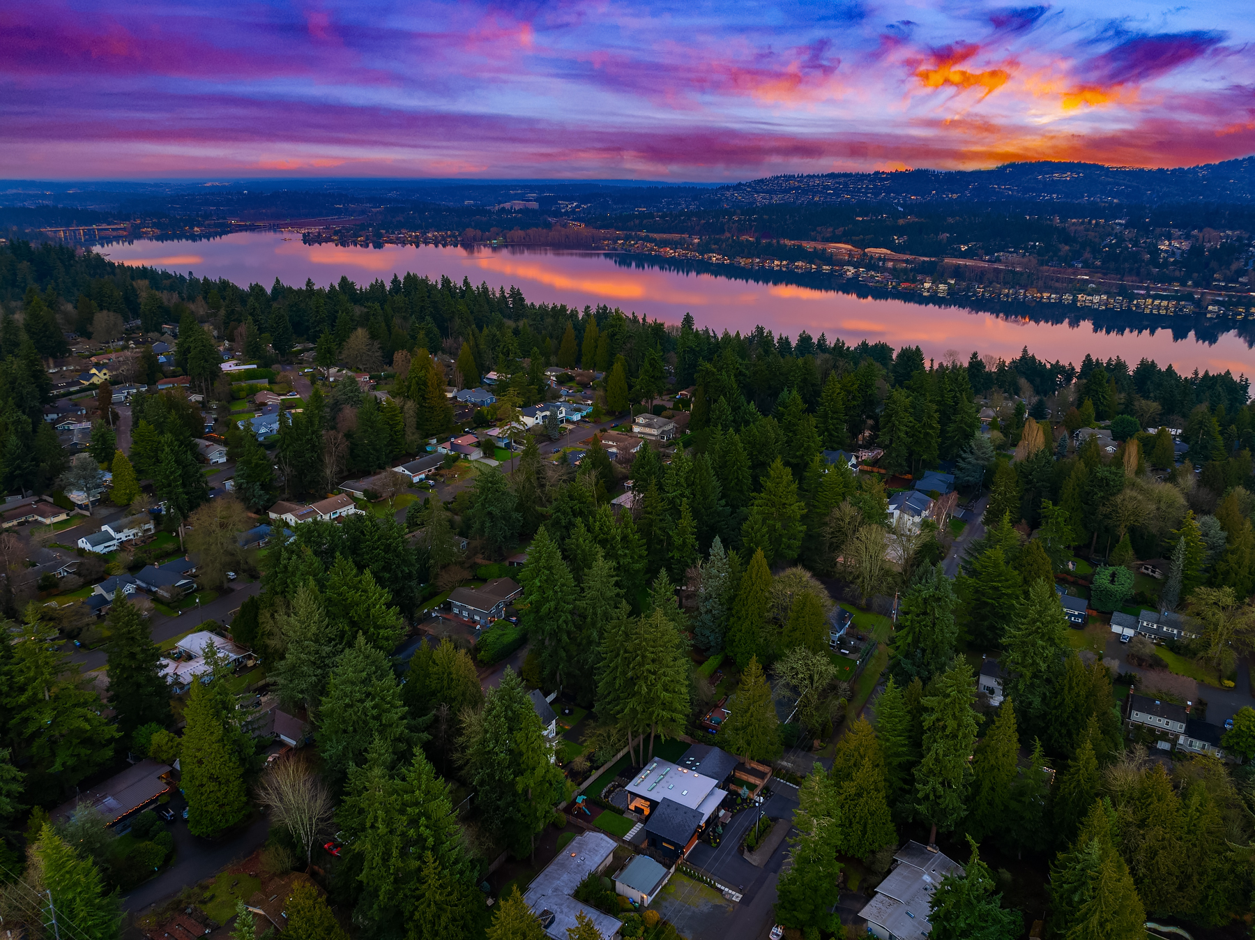 Aerial view of a suburban neighborhood surrounded by dense trees, with a large lake reflecting a colorful sunset sky of pink, purple, and orange hues in the background.