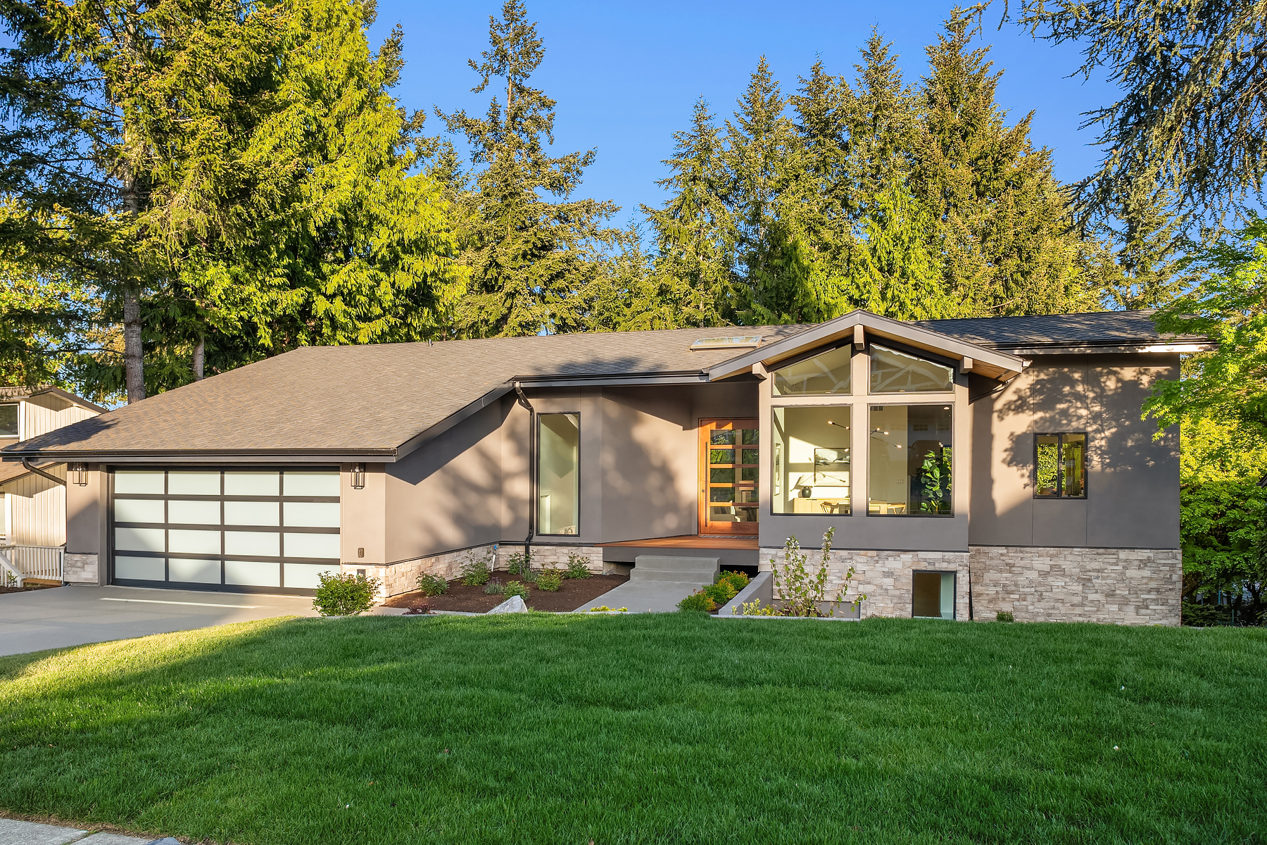 Modern single-story house with large windows, a sloped roof, a two-car garage, and a well-maintained lawn, surrounded by tall evergreen trees under a clear blue sky.