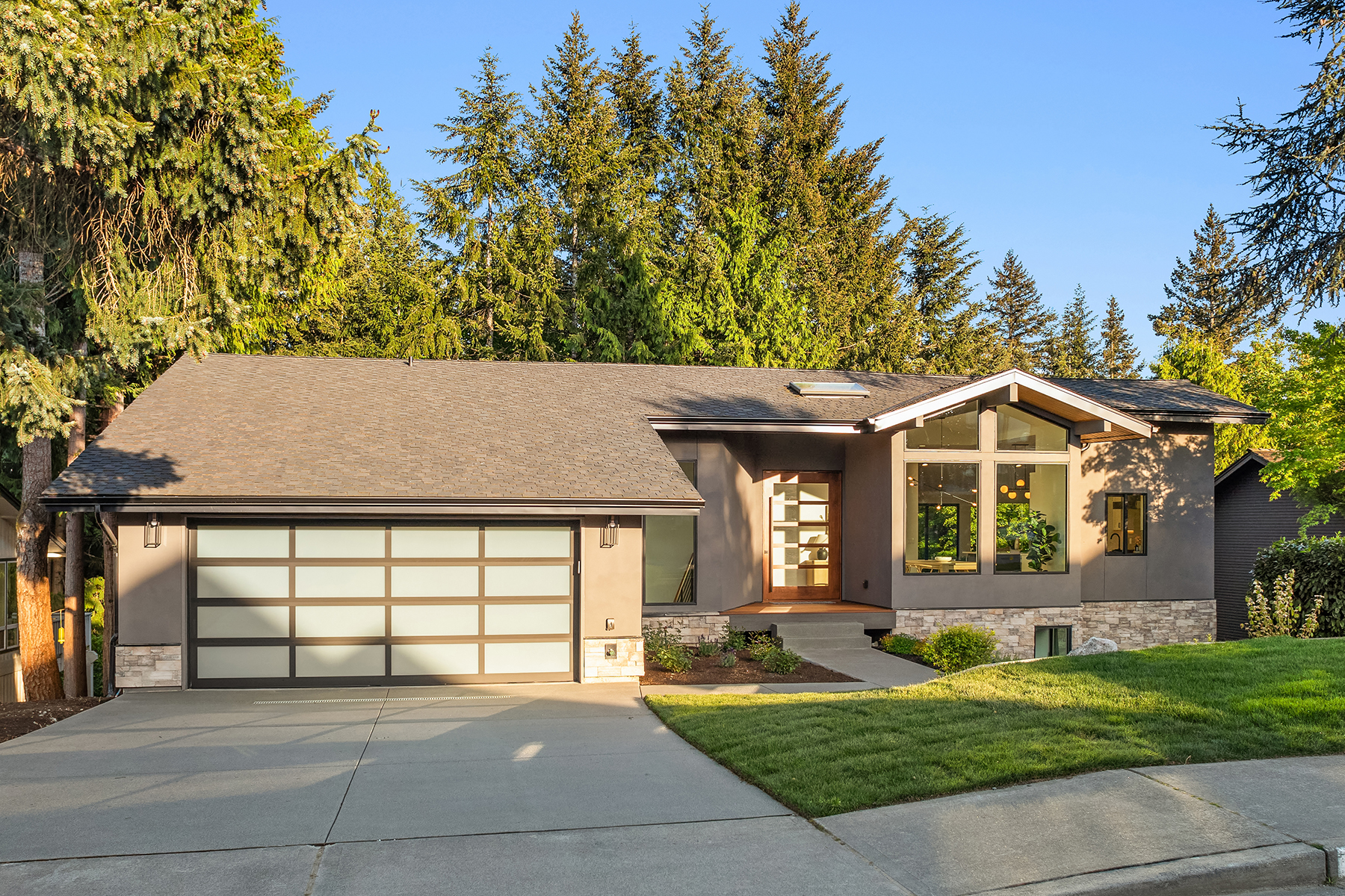Modern single-story house with gray siding, large windows, and a frosted glass garage door. Well-maintained lawn and driveway, surrounded by tall evergreen trees under a clear blue sky.