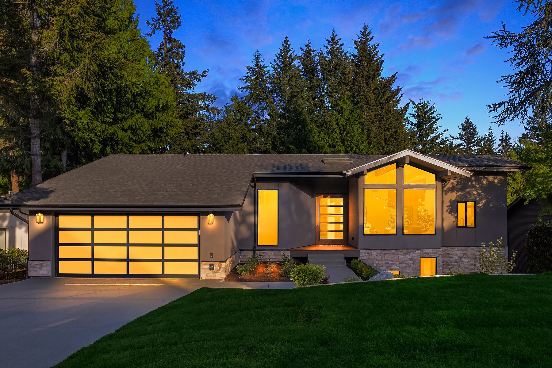 A modern single-story house with large windows glowing with warm light at dusk, a double garage, gray exterior, and surrounded by tall trees and green lawn.